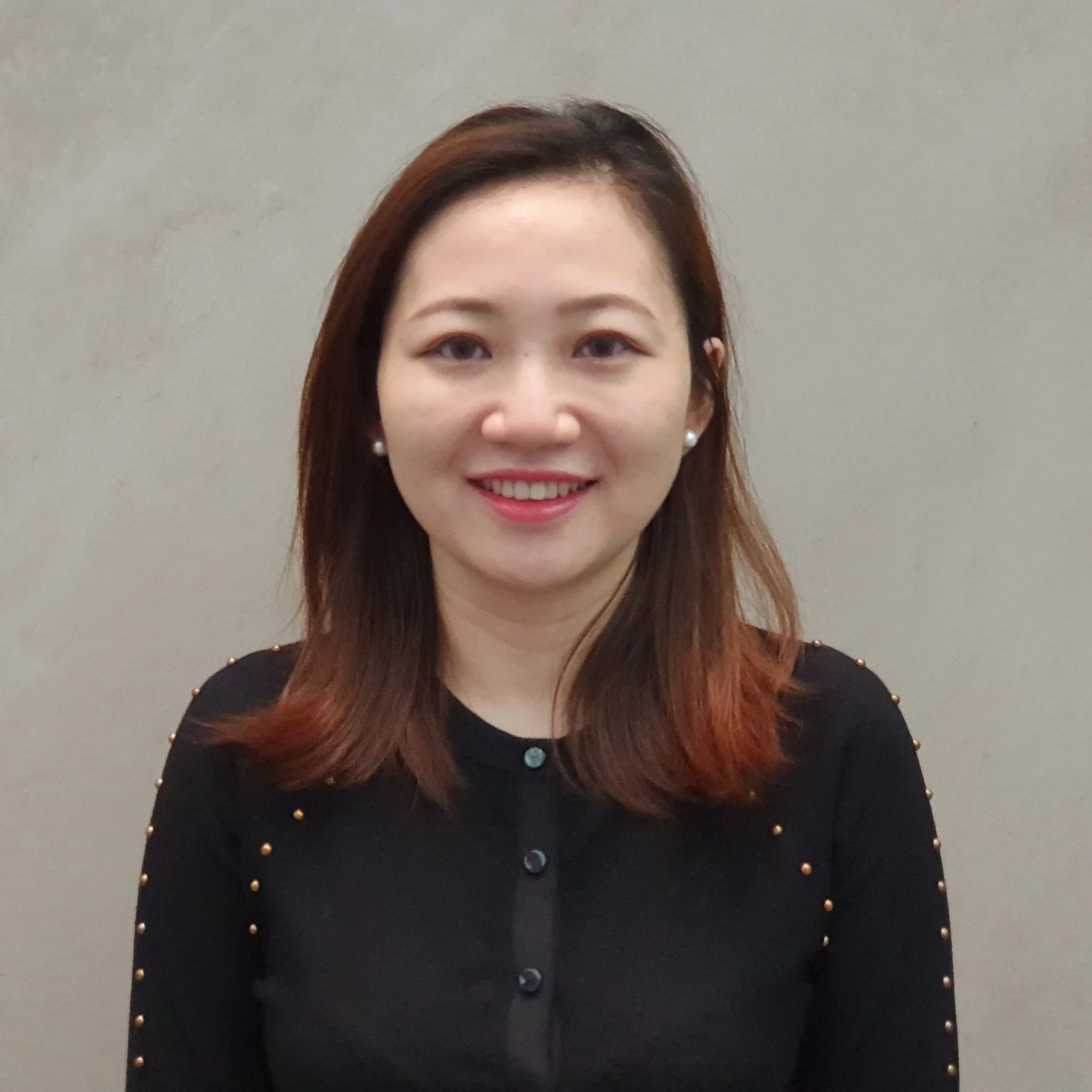 A woman with long reddish-brown hair and pearl earrings smiling in front of a plain gray wall, wearing a black top with gold studded details.