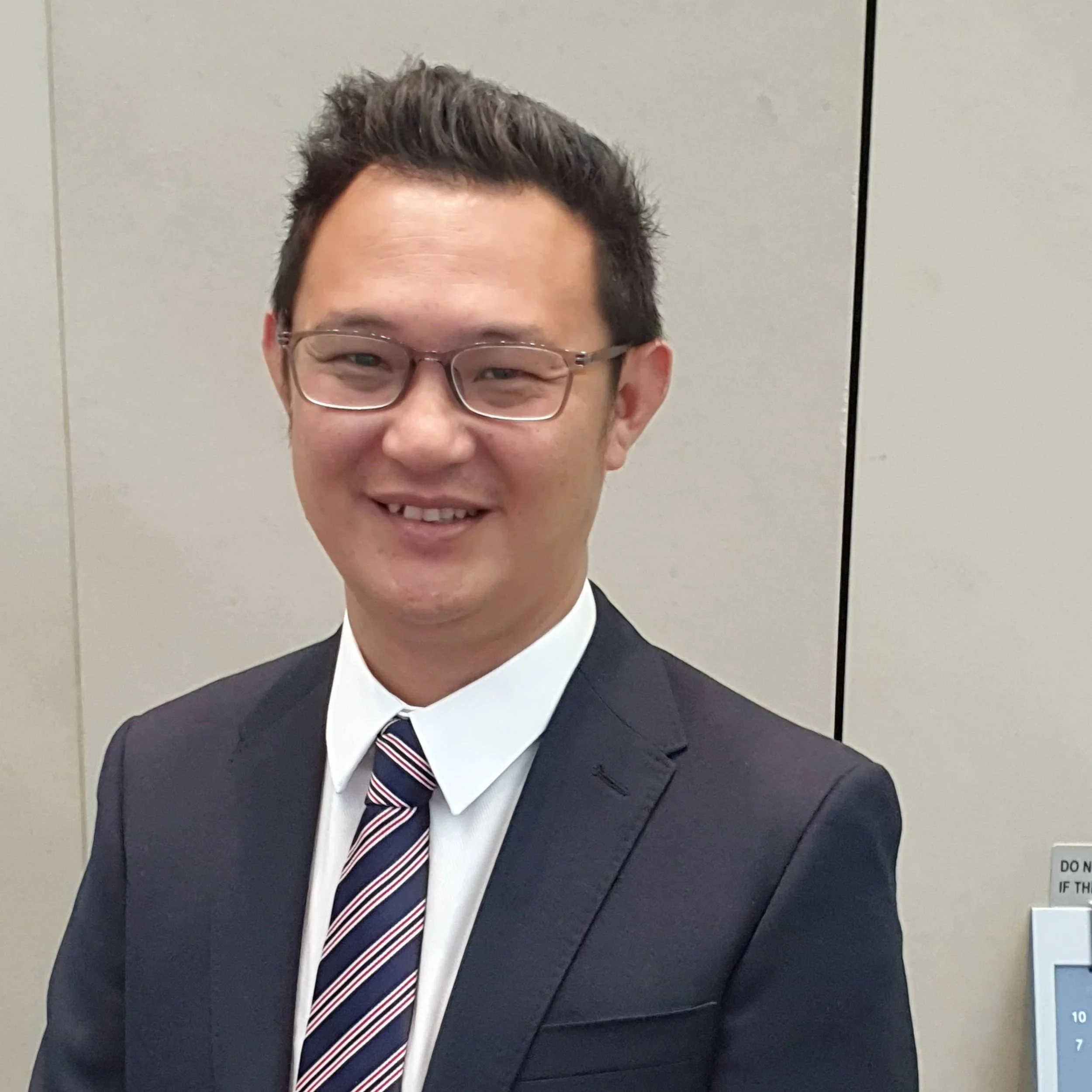 Young man smiling wearing glasses, dark suit, white shirt, striped tie, standing against a light-colored wall with a height measurement chart and a sign reading 'DO NOT' partially visible.