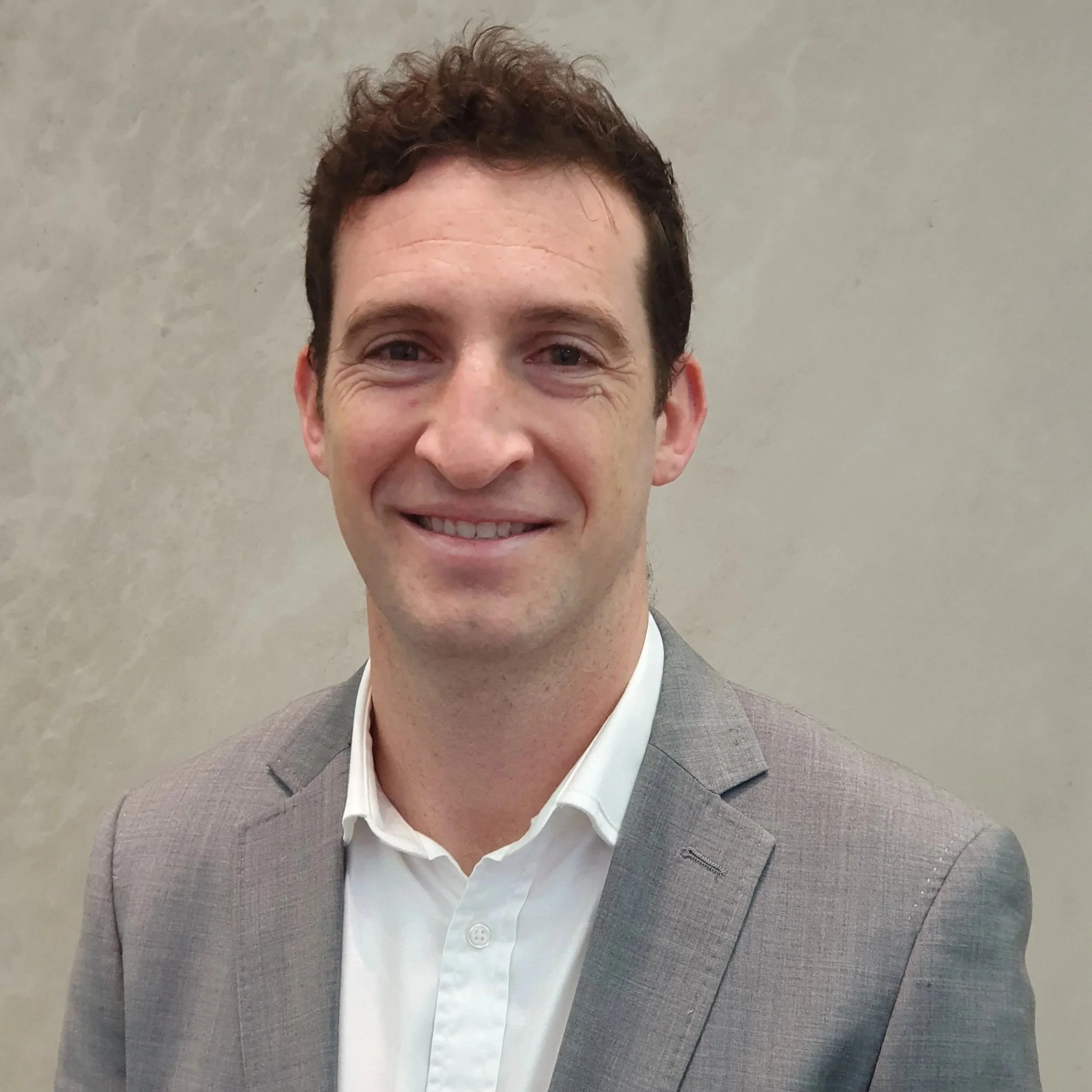 Close-up of a smiling man wearing a light gray blazer and white shirt, standing against a plain neutral background.