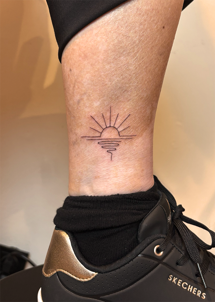 Een enkel met een eenvoudige tattoo van een zonsondergang boven de oceaan, met stil water en stralen die de zon uitstralen.