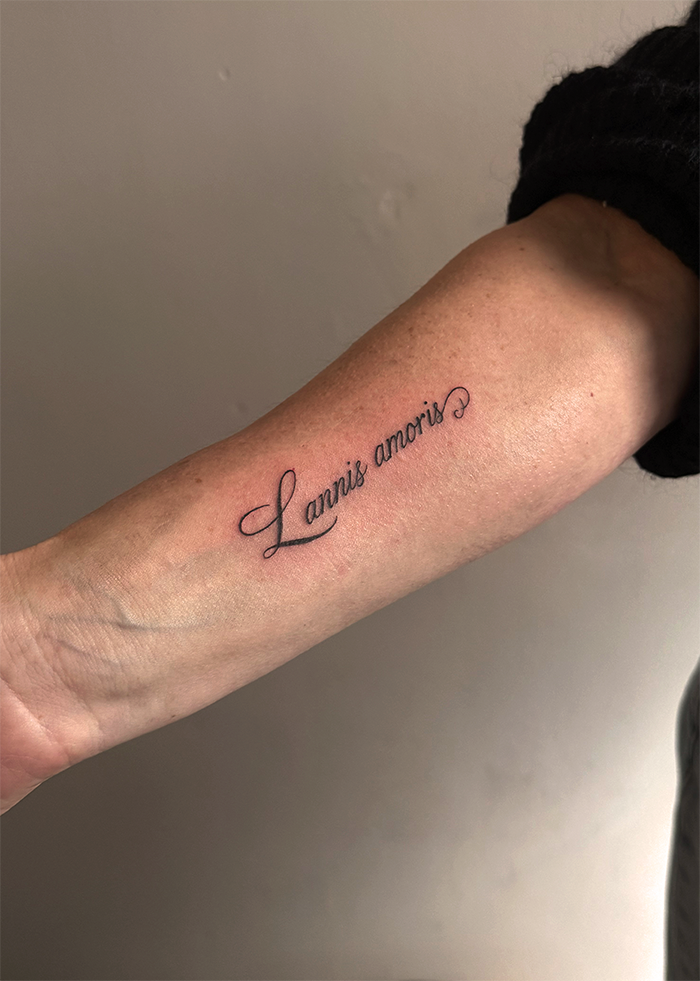 Tattoo op de binnenkant van een onderarm dat zegt 'Lanis amoris' in sierlijke zwarte letters.