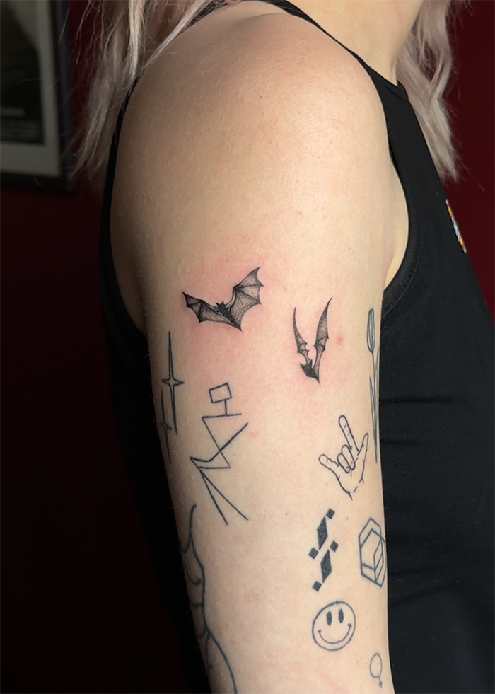 Tattoo van drie vleermuizen op de bovenarm, naast andere kleine tattoo's zoals een handgebaar, klok, smiley en geometrische vormen.