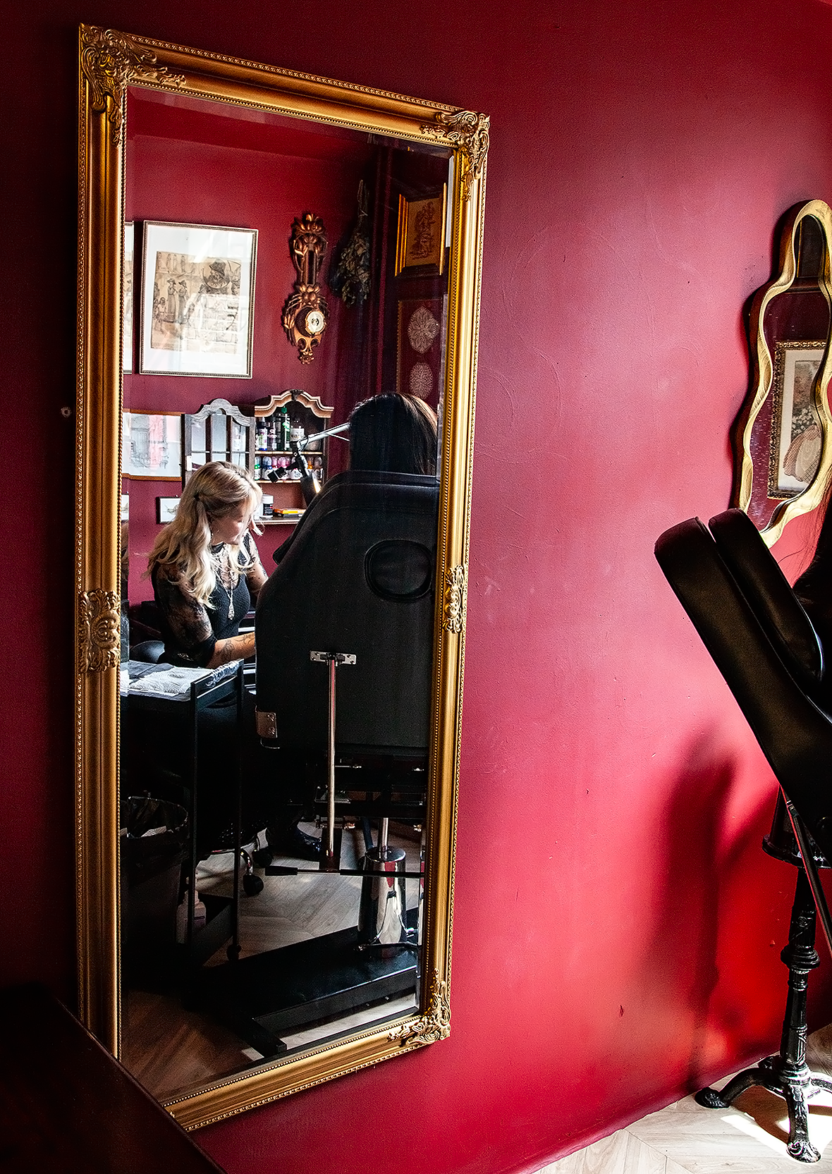 Een persoon wordt in een beautysalon of kapsalon verzorgd, gezien via een grote spiegel met een gouden kader. De kamer heeft roodkleurige muren en heeft diverse decoraties, zoals een ouderwetse klok en kunst aan de muur. Een kaptafel en ander salonmateriaal zijn zichtbaar.
