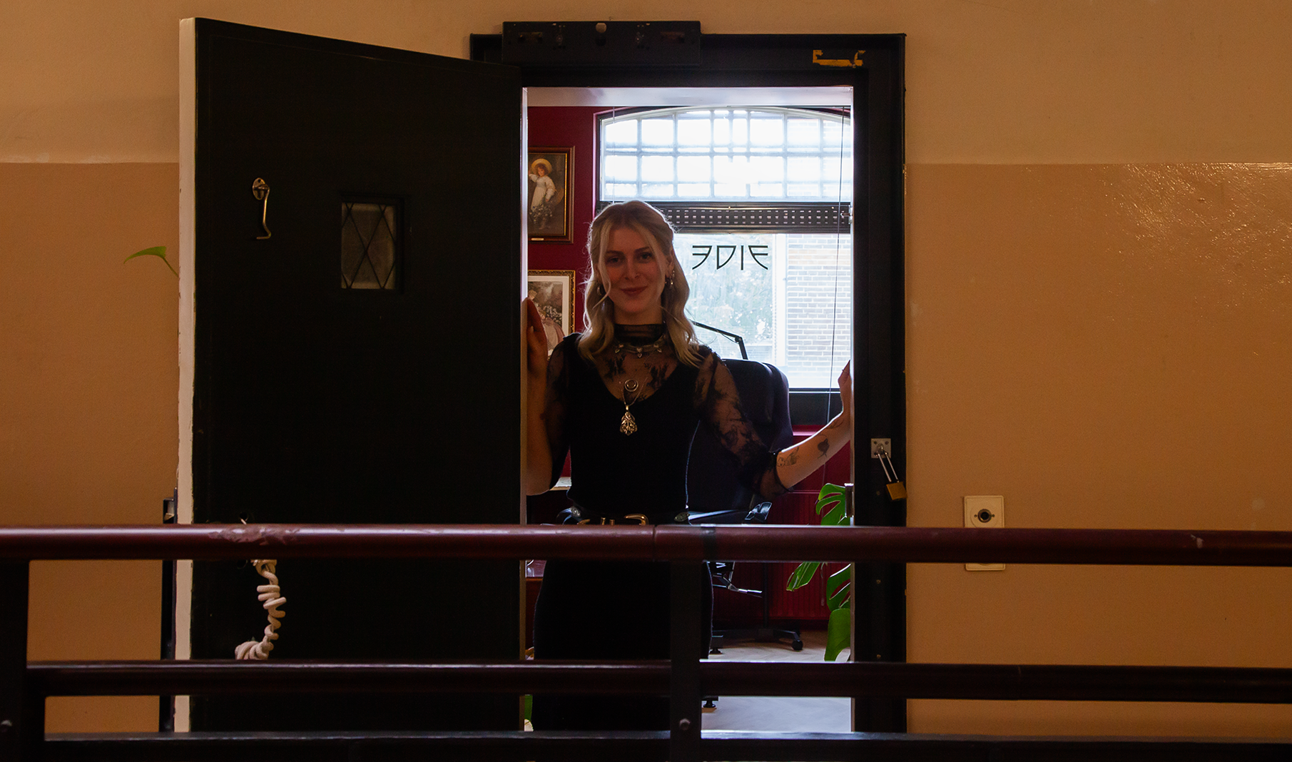 Een vrouw die door een open deur naar buiten kijkt in een binnenruimte, met twee schilderijen aan de muur en een grote raam in de achtergrond.