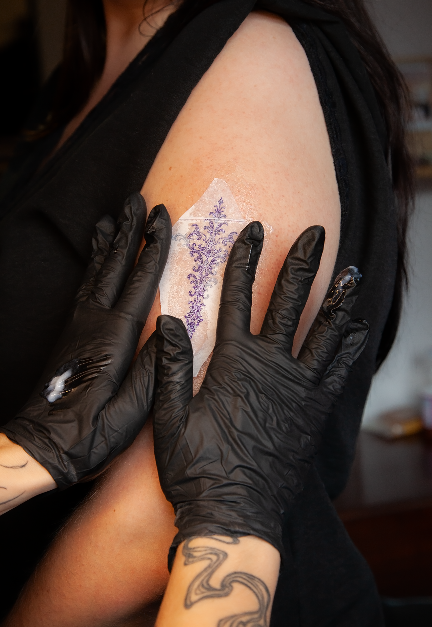 Persoon krijgt een tattoo gezet op de bovenarm. De tattoo is nog niet volledig afgewerkt en de persoon draagt zwarte handschoenen.