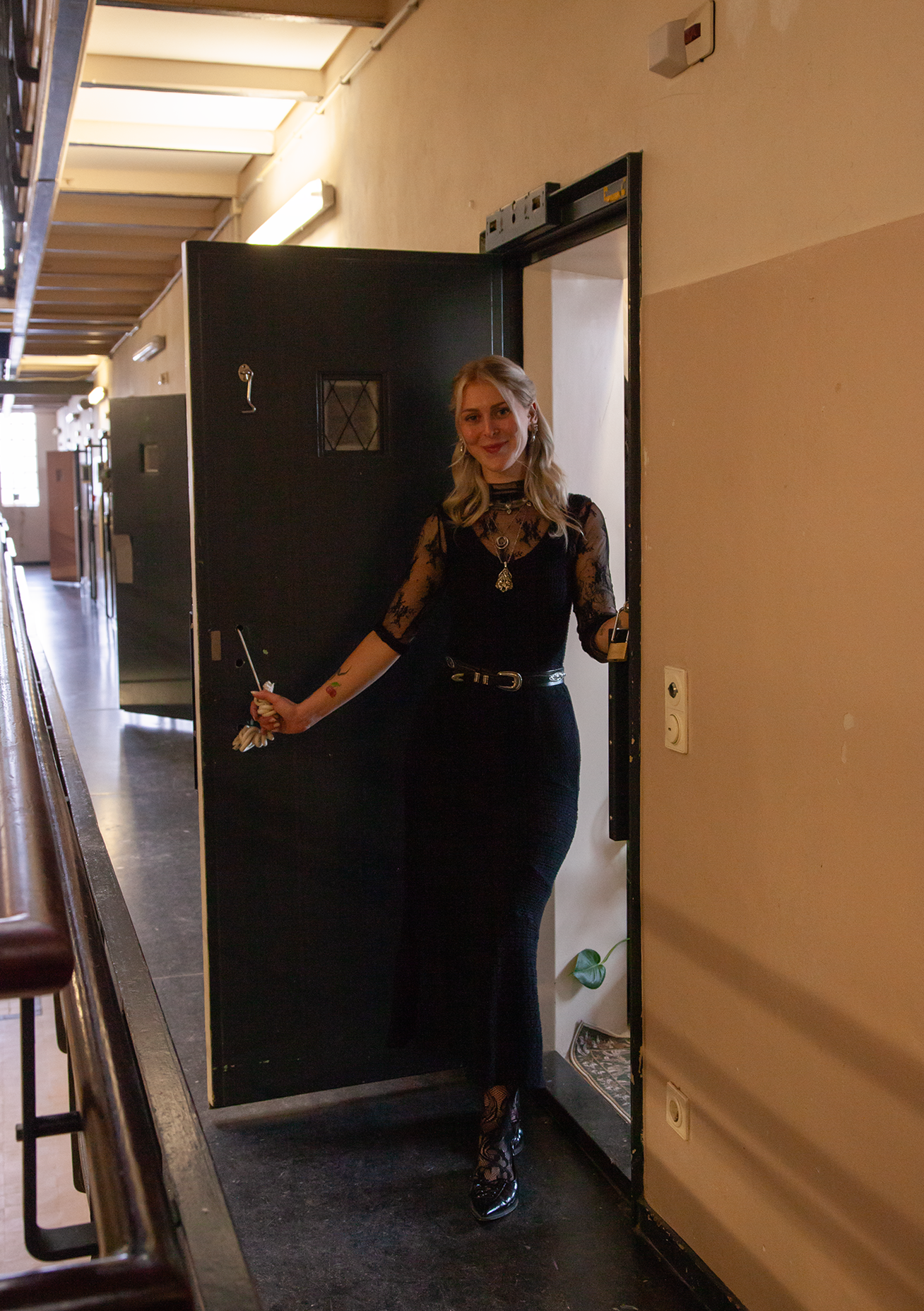 Een vrouw staat in een deuropening in een gang, glimlacht naar de camera. Ze draagt een zwarte jurk met doorzichtige mouwen en lace details, met een lange rok en hoge hakken. Ze houdt een paraplu vast in haar rechterhand.