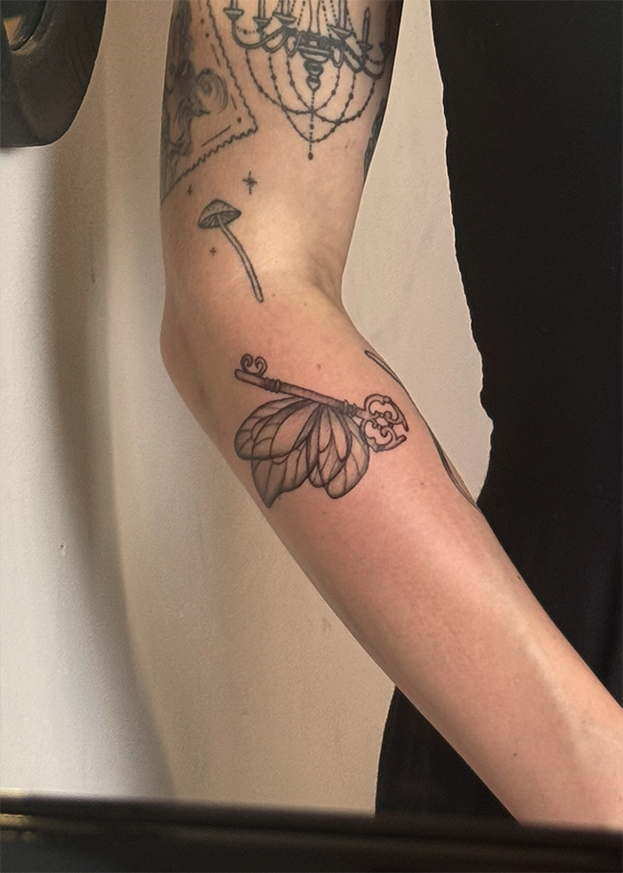 Tatoeage van een sleutel met vleugels op de onderarm, een parasol en een kroon in de buurt.