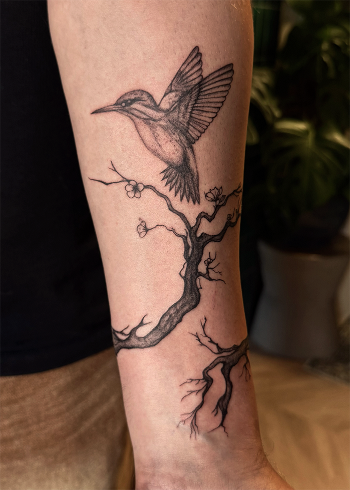 Tattoo van een hagedis die uit een boomtop springt, getekend met zwarte lijnen, op de binnenkant van de onderarm.