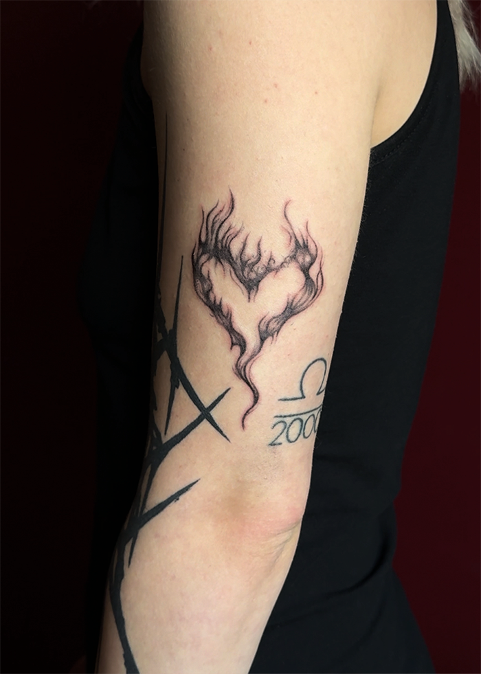 Tattoo van een hart gevormd door vlammen op de bovenarm naast een teken en het jaar 200