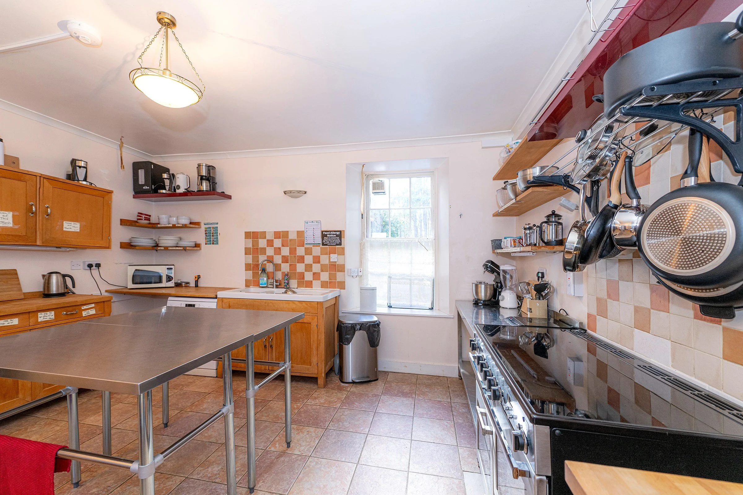 A kitchen with wooden cabinets, open shelves, and a tiled backsplash. There are various kitchen appliances, including a microwave, coffee makers, and a stove. Kitchen tools hang above the stove. A window with an air conditioning unit is visible, and a door leads outside. The room has a tiled floor and a ceiling light fixture.