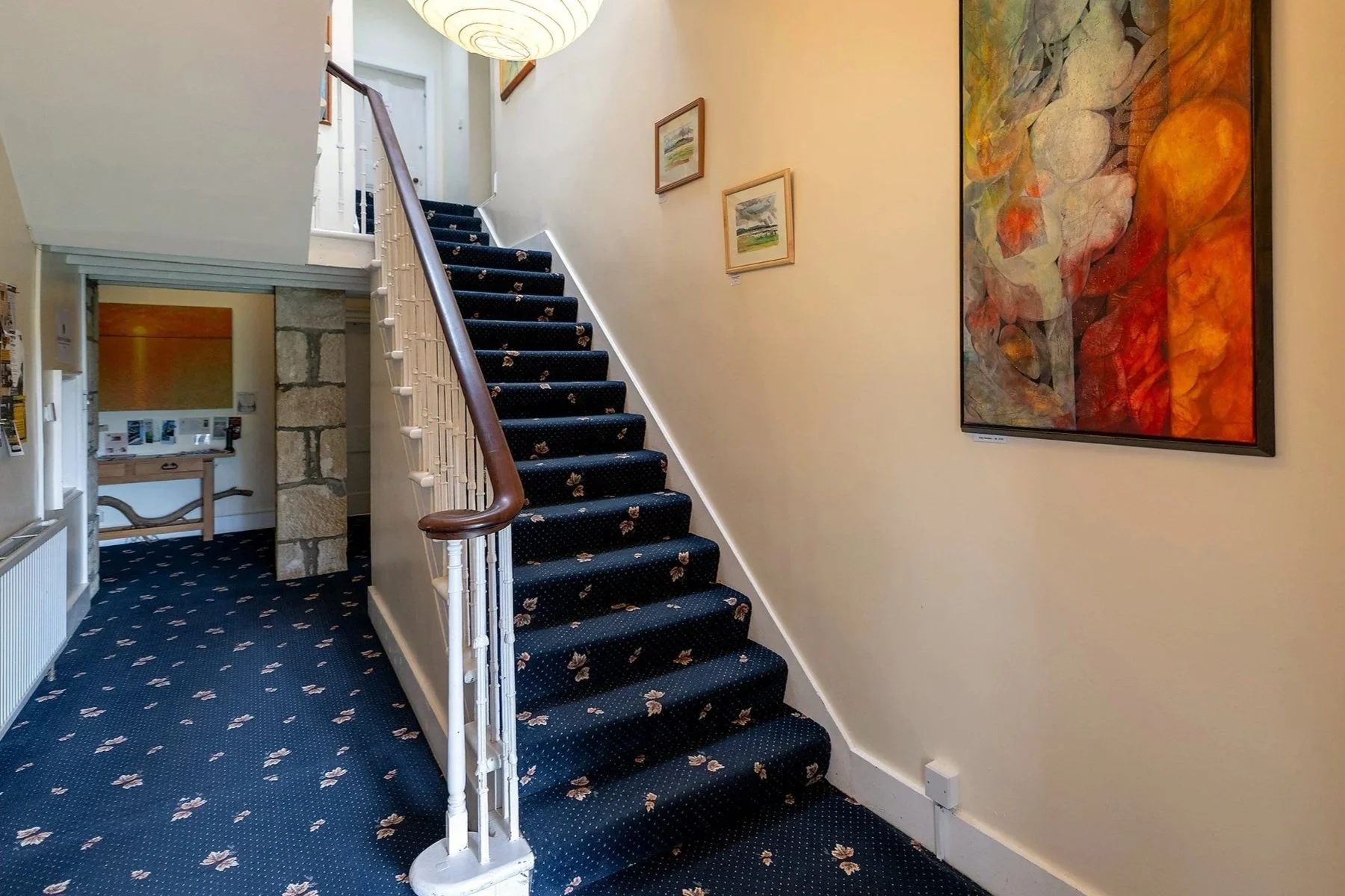 Interior view of a staircase with dark blue carpeted stairs decorated with floral patterns, a wooden handrail, and white railings. The wall has framed artwork, and a large abstract painting is visible on the right. The area is well-lit, with a round paper lantern hanging from the ceiling.