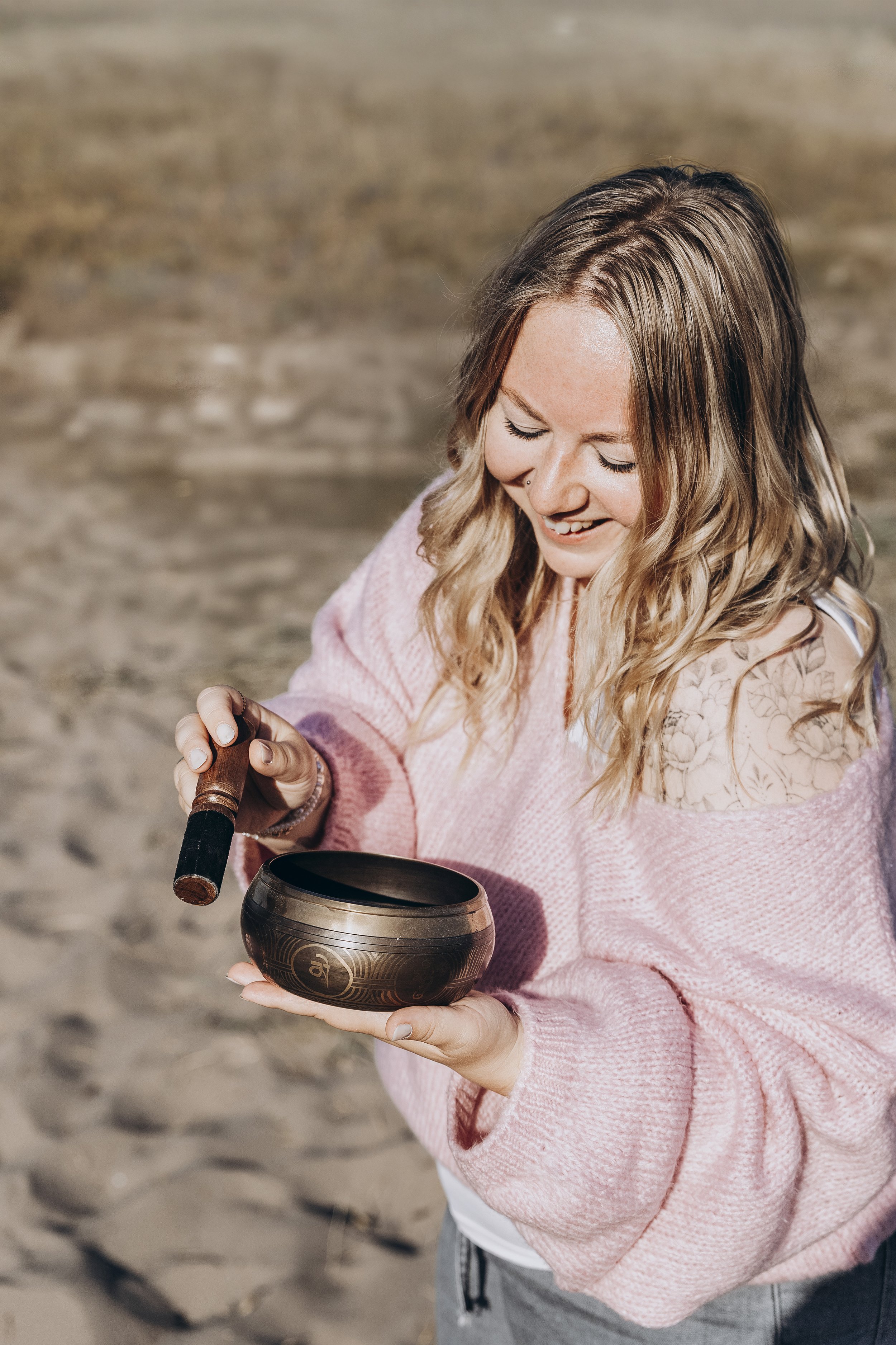 Junge Frau im pinken Pullover hält eine tibetische Klangschale und benutzt einen Schlägel, während sie am Strand steht.