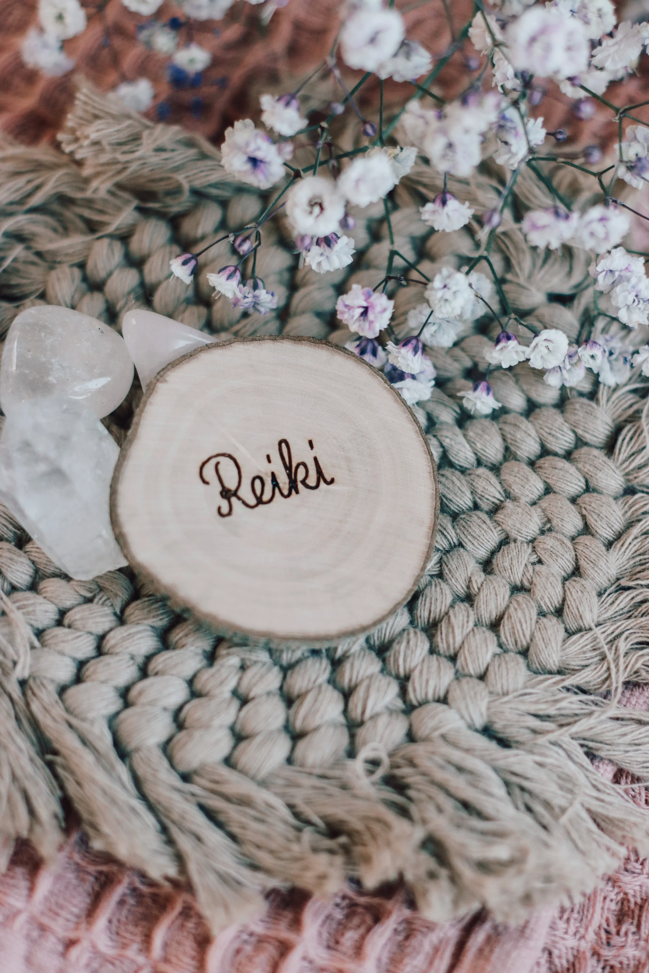 Eine Holzscheibe mit dem Schriftzug 'Reiki' neben weißen Kristallen, auf einem handgefertigten Stick-Teppich mit einer Borte in Rosé-Farben, umgeben von kleinen blühenden Zweigen.