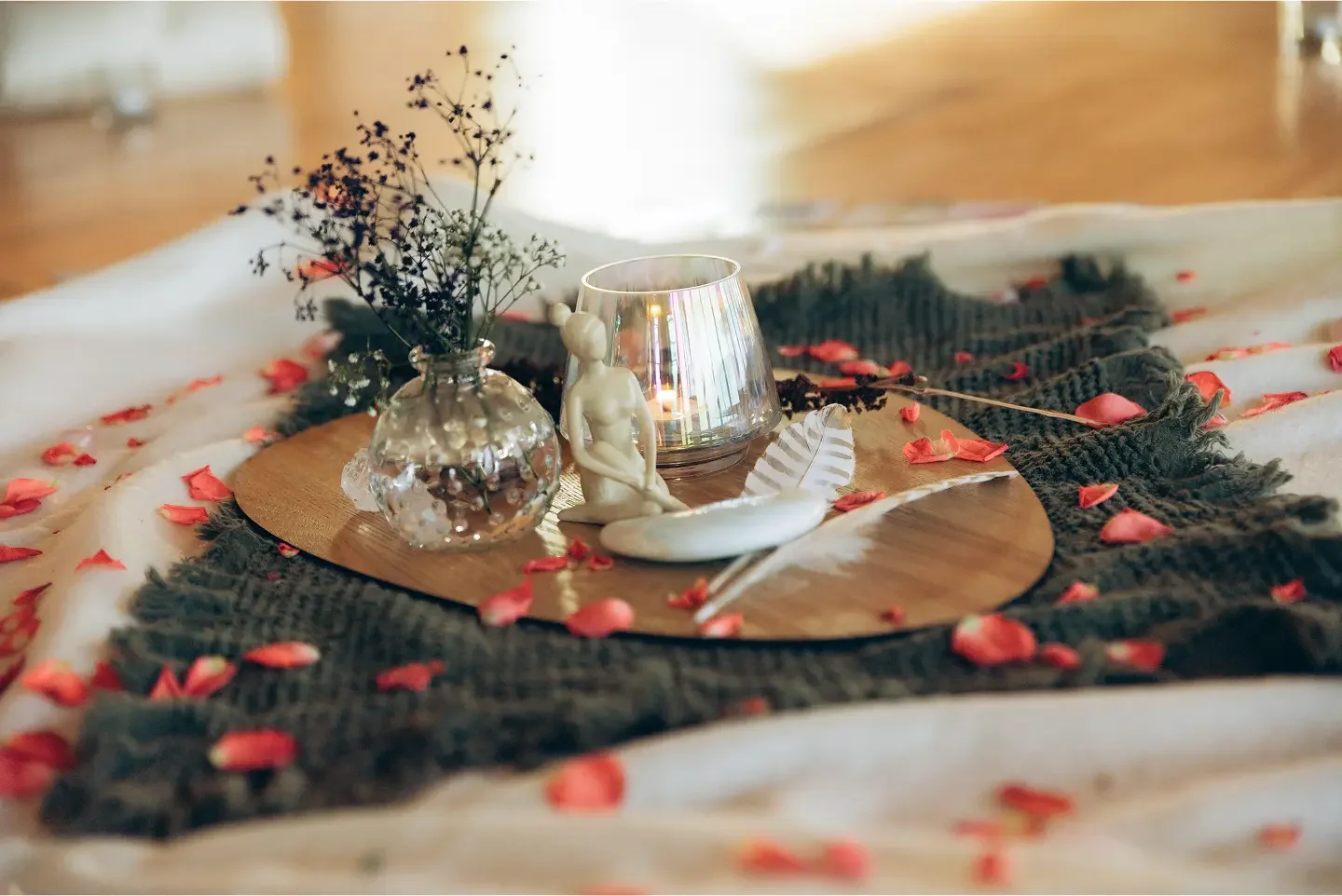 Verschiedene Dekorationselemente auf einem runden Holztablett, darunter eine kleine Skulptur, eine Kerze in einem reflekierenden Glas, eine Vase mit Blumen und eine Feder, umgeben von Rosenblättern auf einem Stoffuntergrund.
