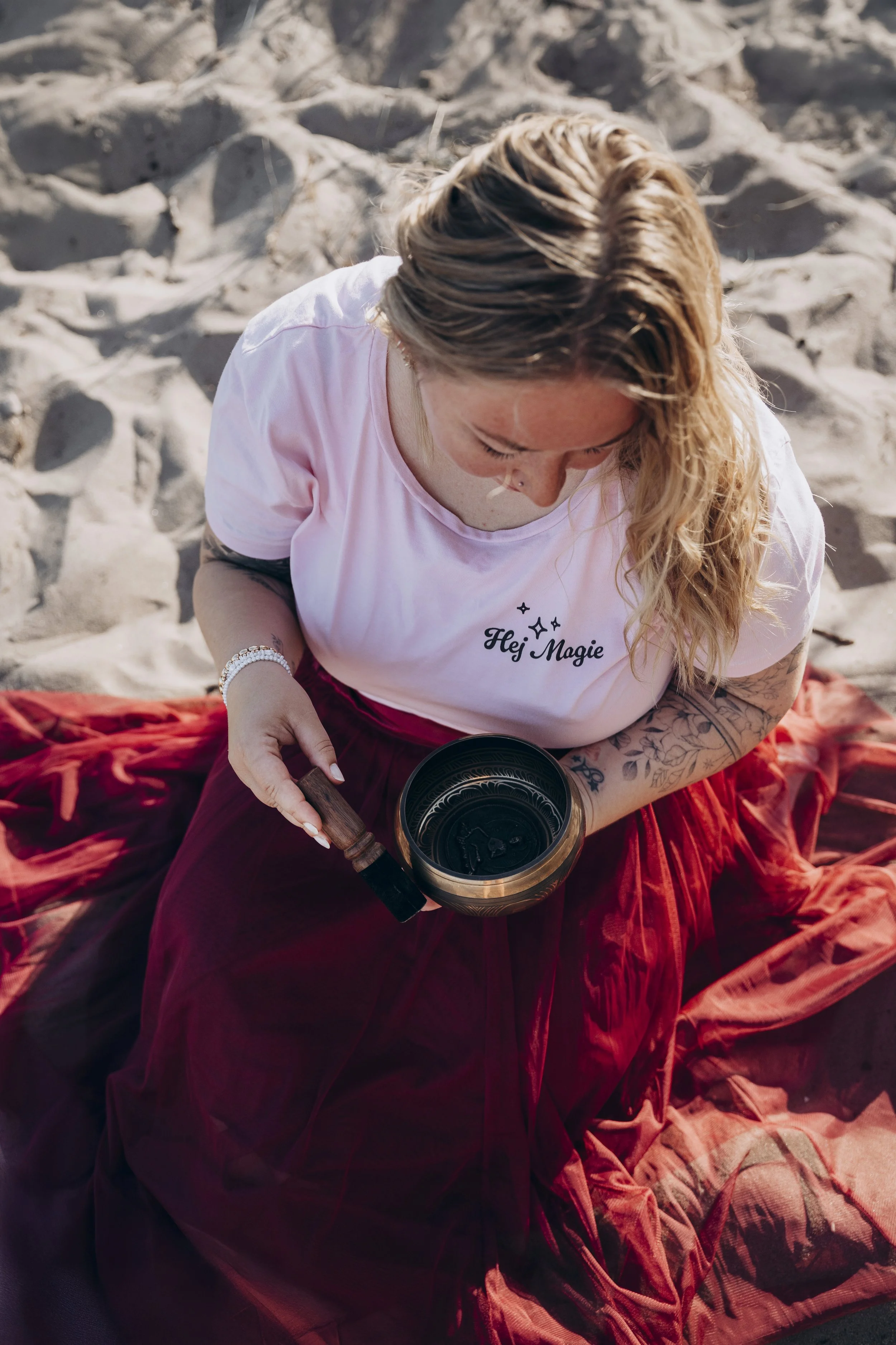 Eine Frau mit langem, gewelltem, blondem Haar sitzt auf einem roten Samtstoff auf einem Sandstrand, hält eine Walnussmühle und eine Schale. Sie trägt ein hellrosafarbenes T-Shirt mit der Aufschrift "Hej Magie" und Tattoos am linken Arm.