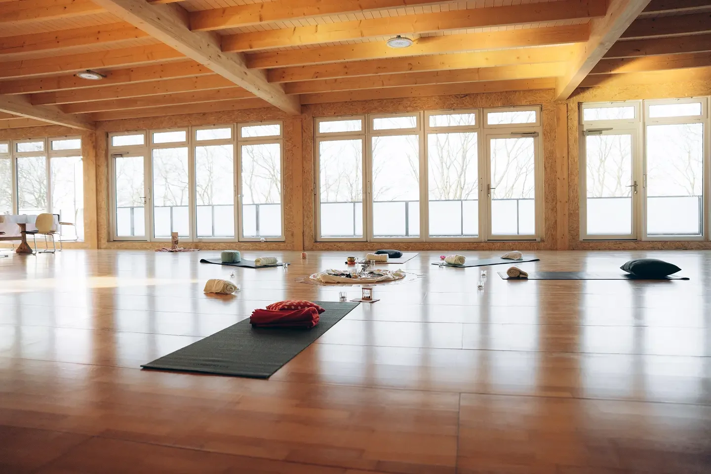 Ein heller, leerer Yoga-Raum mit Holzdecken, großen Fenstern und Yogamatten, die mit Decken, Kissen und Kerzen vorbereitet sind.