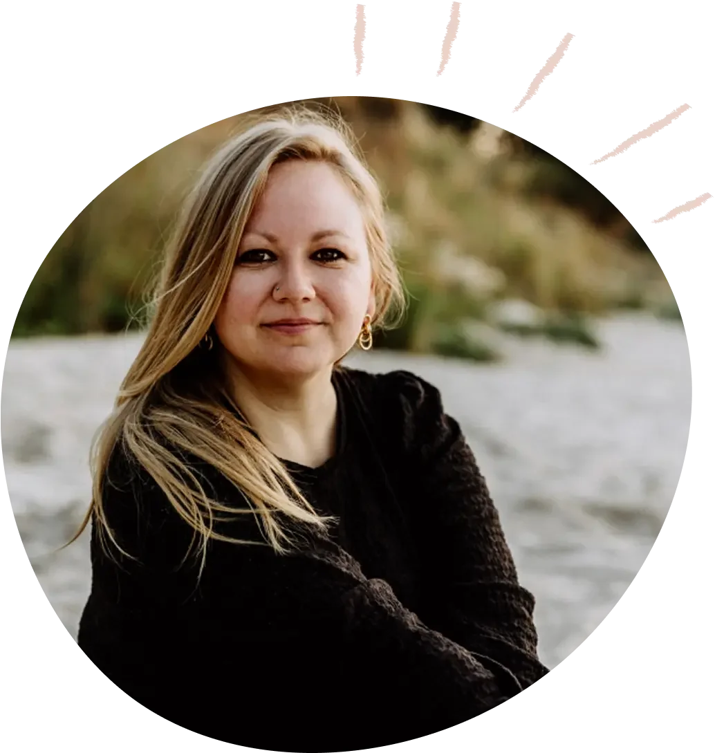 Porträt einer Frau mit blondem Haar, die lockere schwarze Kleidung trägt, vor einem Fluss im Freien, bei Sonnenuntergang, mit Bäumen im Hintergrund.