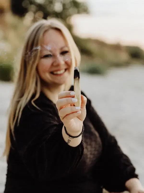 Junge Frau am Wasser beim Lagerfeuer, hält eine brennende Kohle in der Hand, lacht, trägt schwarze Kleidung und hat lange blonde Haare.