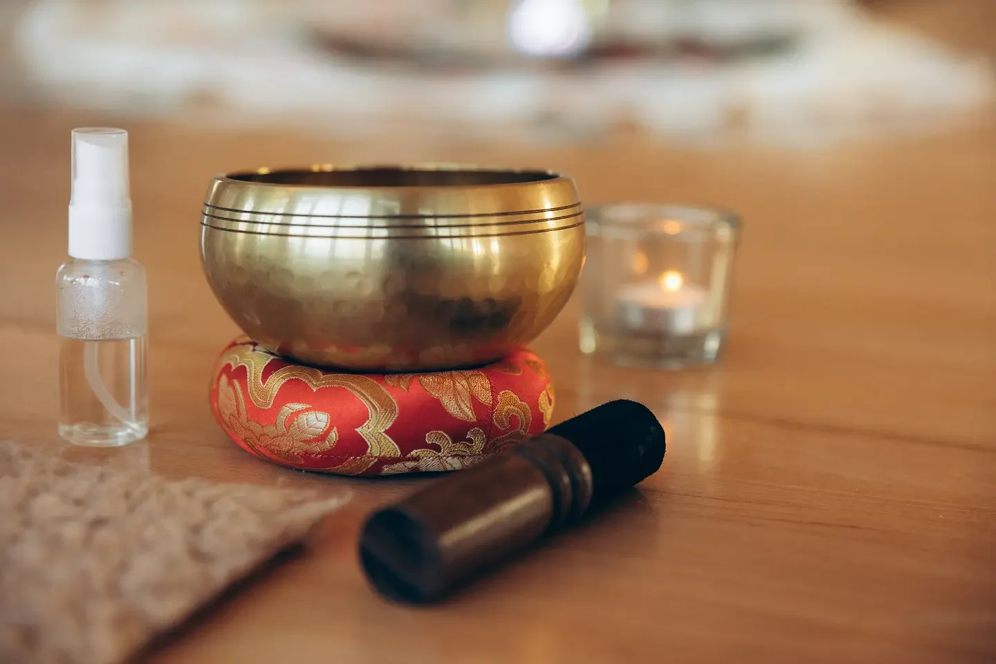 Ein Klangschale, auf einer rot-goldenen Kissen, ein kleines Spray, eine Kerze und ein Massageholz liegen auf einem Holztisch, typisch für eine Entspannungs- oder Meditationssitzung.