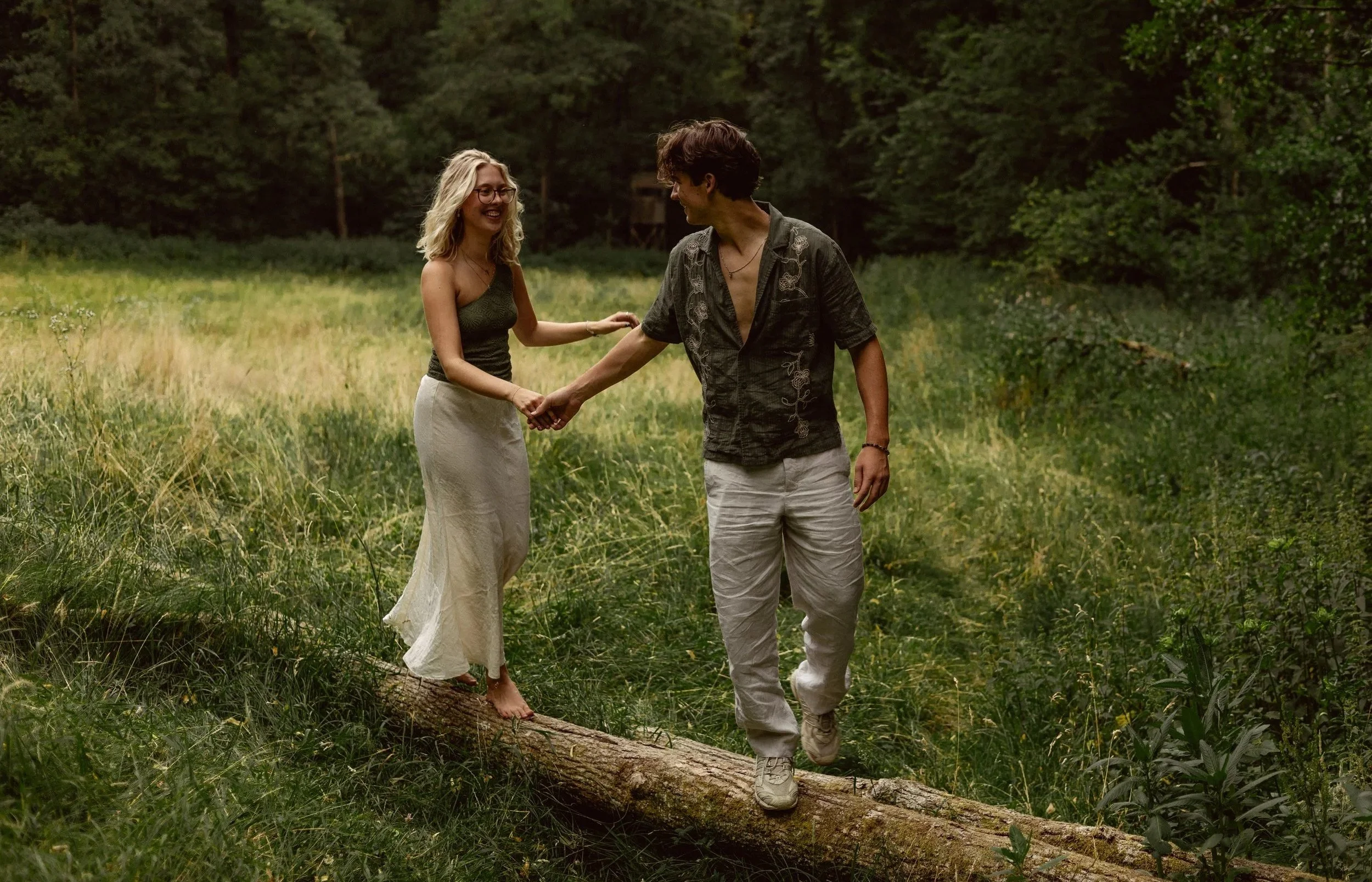 A young couple holding hands and walking on a fallen tree in a lush green forest, smiling at each other.