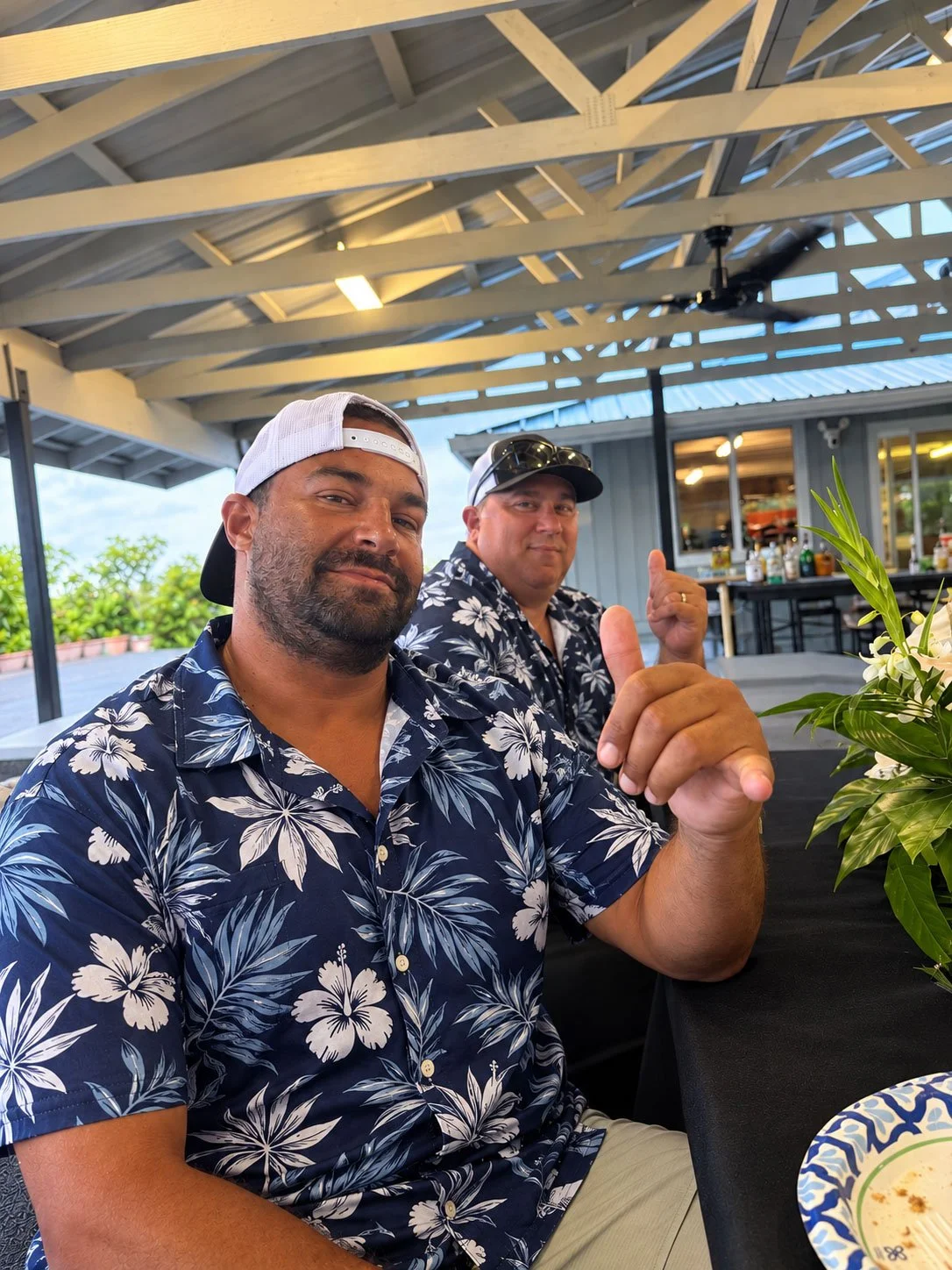 Two men sitting at a table, wearing matching floral shirts, with one giving an OK sign and the other with sunglasses on. They are in an outdoor area with a wooden roof and a bar in the background, and a plant is in the foreground.