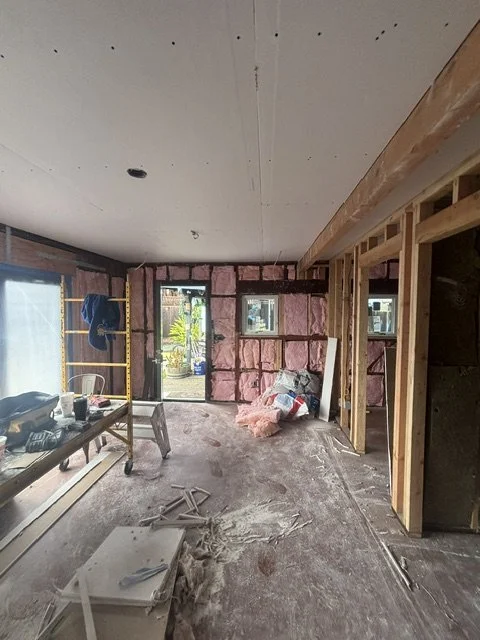 Interior of a room under construction with exposed framework, pink insulation, construction tools, and materials, with a door leading outside.