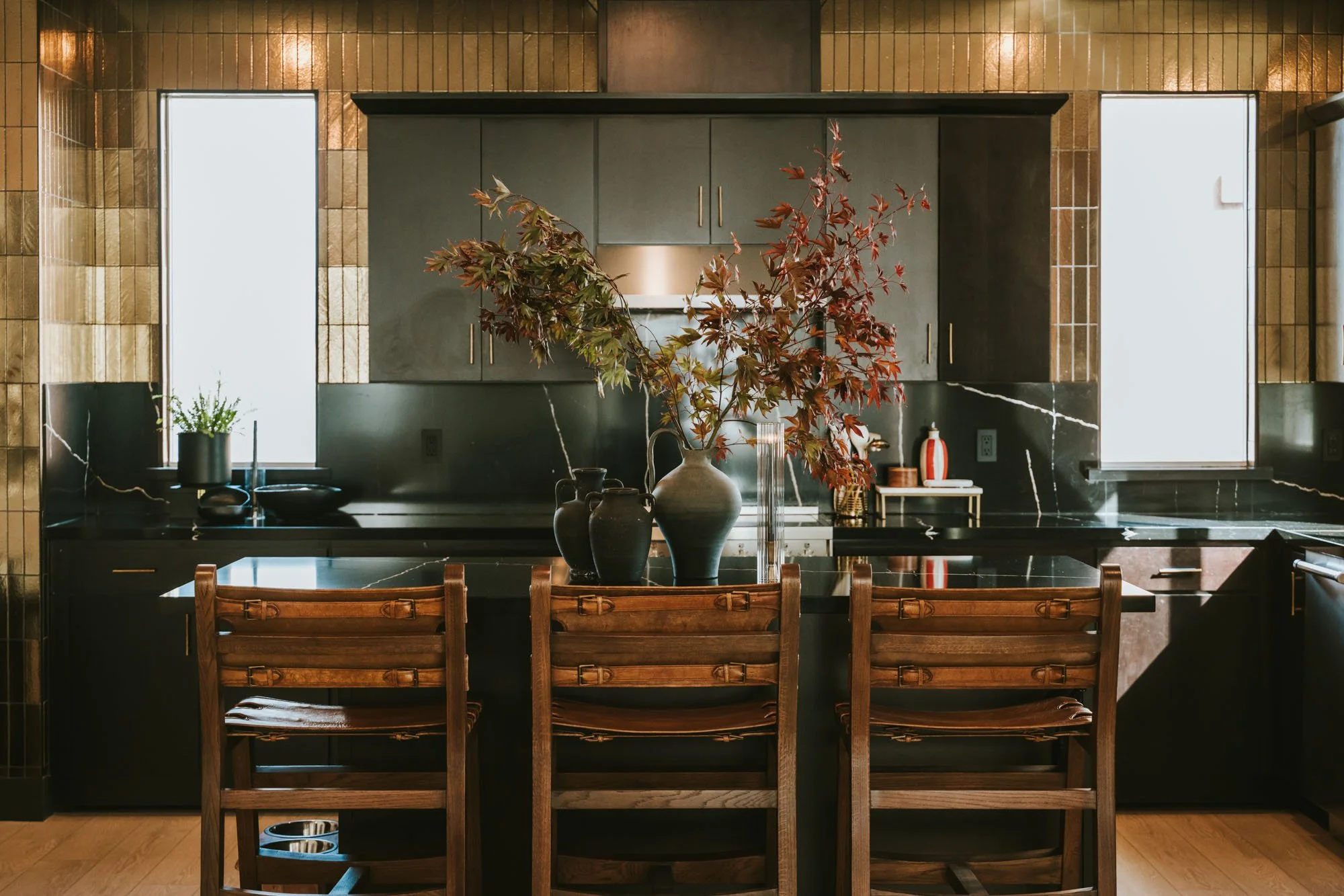 A modern kitchen with a black marble island, a large vase with red and green foliage, wooden chairs, and blonde wood flooring. Two tall windows with frosted glass flank the backsplash.