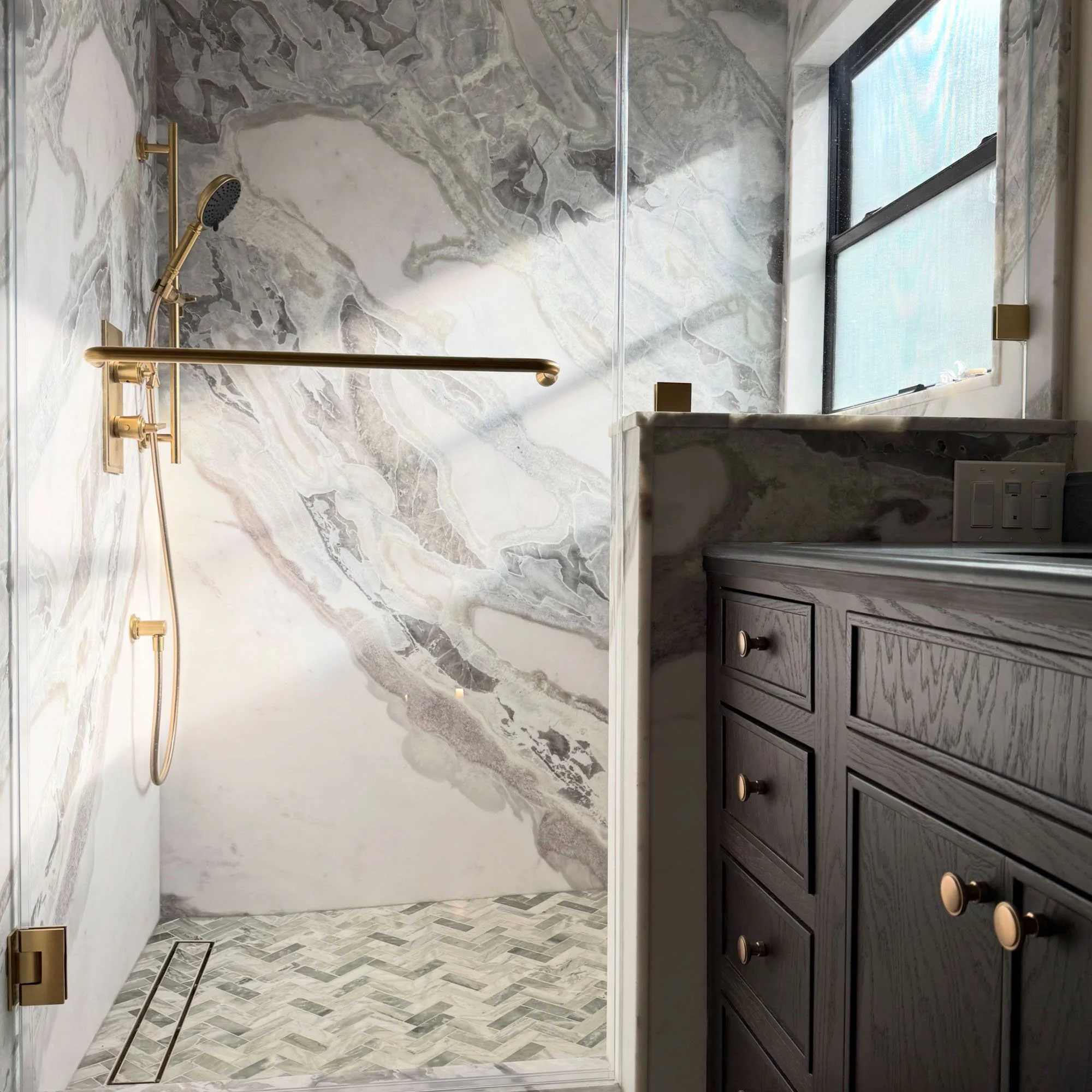 A modern bathroom shower area with marble wall tiles, a gold showerhead and fixtures, a glass shower door, a window with black framing, and a dark wooden vanity with gold knobs.