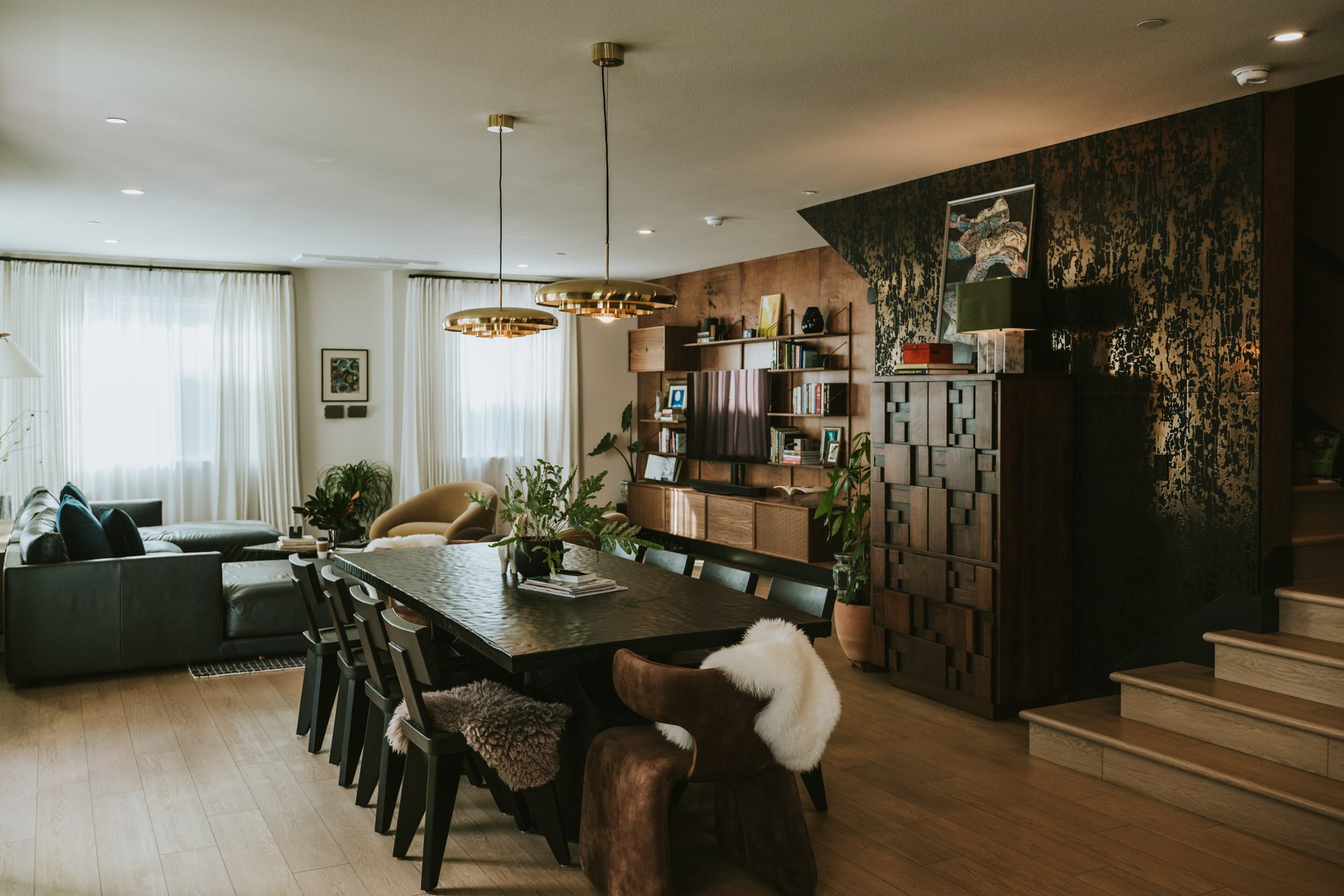 Interior of a modern living and dining area with a long dining table, black chairs, a wood panel wall with shelves, modern pendant lights, a sofa, and curtains covering large windows.