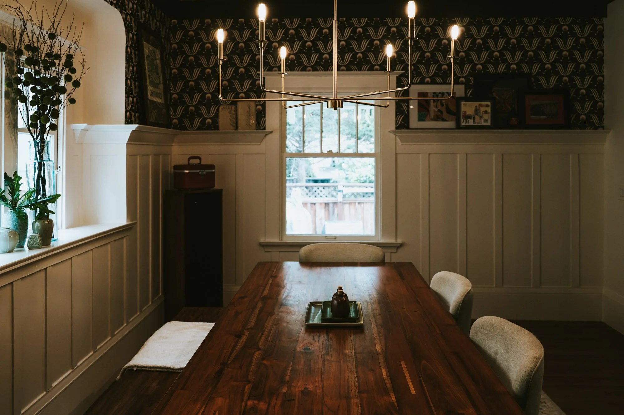 A dining room with a wooden table, beige chairs, a window with a view of a backyard, a chandelier with multiple bulbs, wall art, and decorative plants near the window.