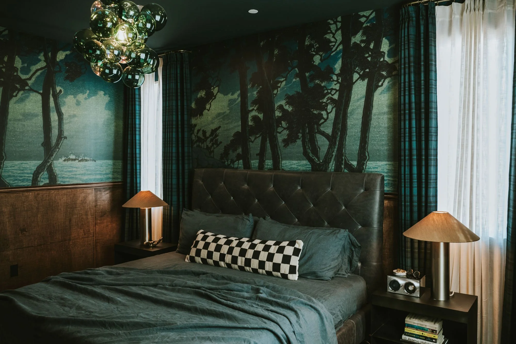 A bedroom with dark green walls featuring a large mural of trees and water. A dark brown tufted leather headboard, gray bedspread, and black-and-white checkered pillow. Two bedside lamps with gold bases and beige lampshades. A stack of books and a sm