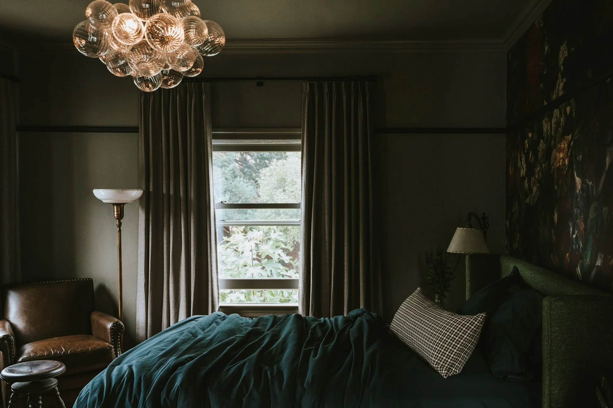 A dimly lit bedroom with a bed covered in dark green bedding, a patterned throw pillow, and a window with sheer curtains allowing some sunlight to enter. There is a modern chandelier overhead, a tall floor lamp, a leather armchair, and floral wallpap