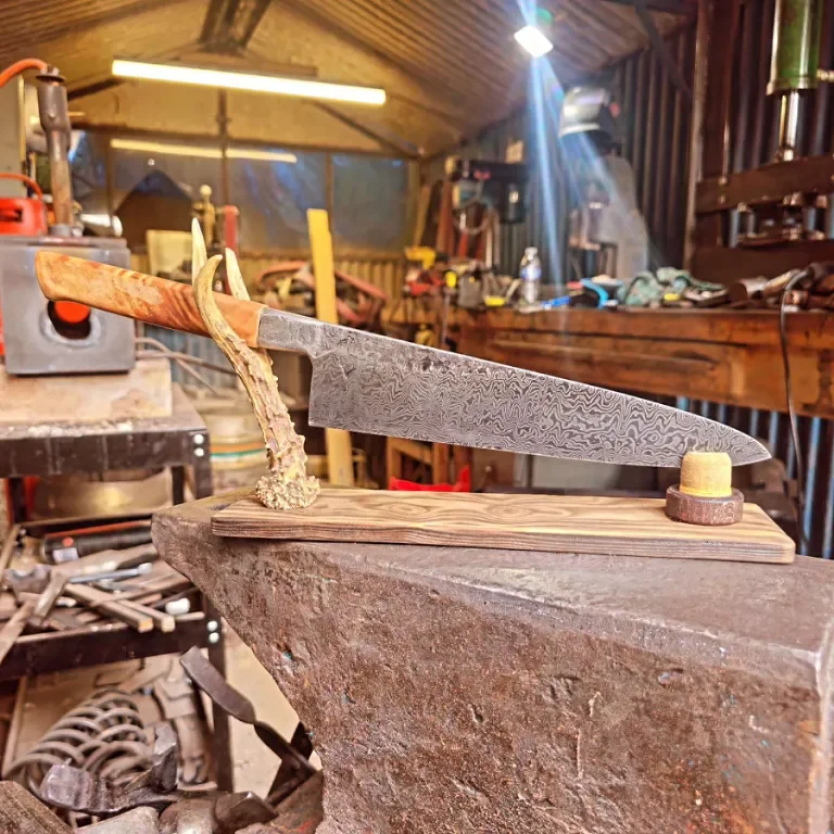 A sharp chef's knife with a Damascus steel pattern on the blade, mounted on a wooden stand in a woodworking or blacksmithing workshop.