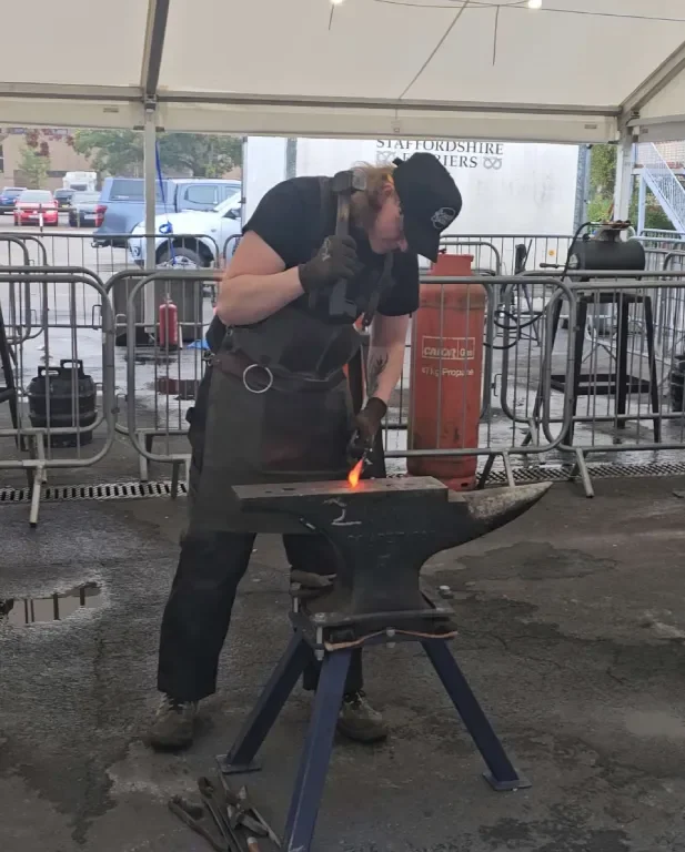 Blacksmith working at an outdoor event under a tent, using a small anvil and a torch to heat and shape metal.