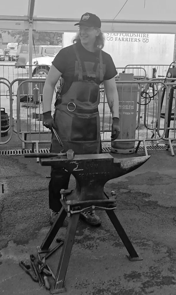 Black and white photo of a smiling woman wearing a helmet, apron, and gloves, working at a blacksmithing station with an anvil and tools inside a tent. Behind her, there are metal barriers and trucks outside.