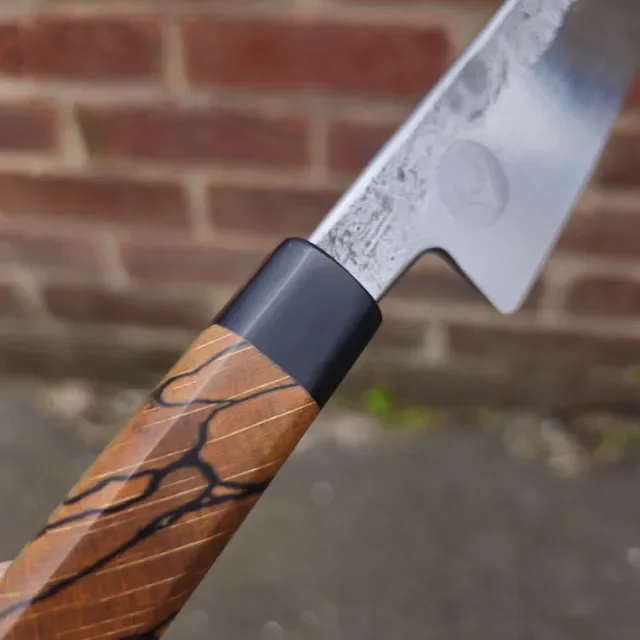 Close-up of a kitchen knife with a wooden handle and a steel blade, held outdoors against a brick wall background.