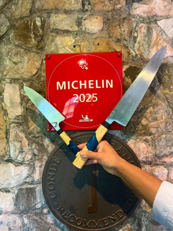 A person's hand is holding two large chef knives crossed in front of a stone wall. Behind the knives, there is a red plaque with the Michelin logo and the year 2025. Below directly is a black circular plaque with the words "CONDE NAST RECOMMEND" around the edge.