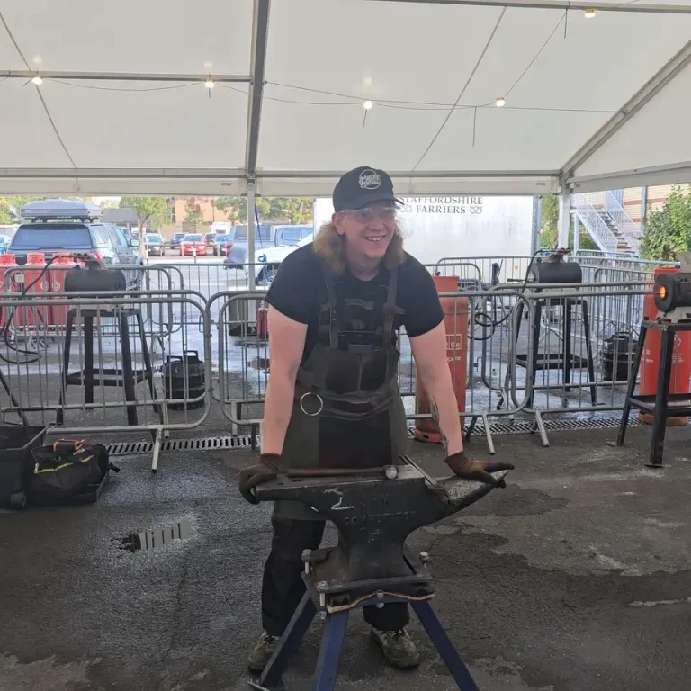 Smiling blacksmith working at an anvil under a tent at an outdoor event.