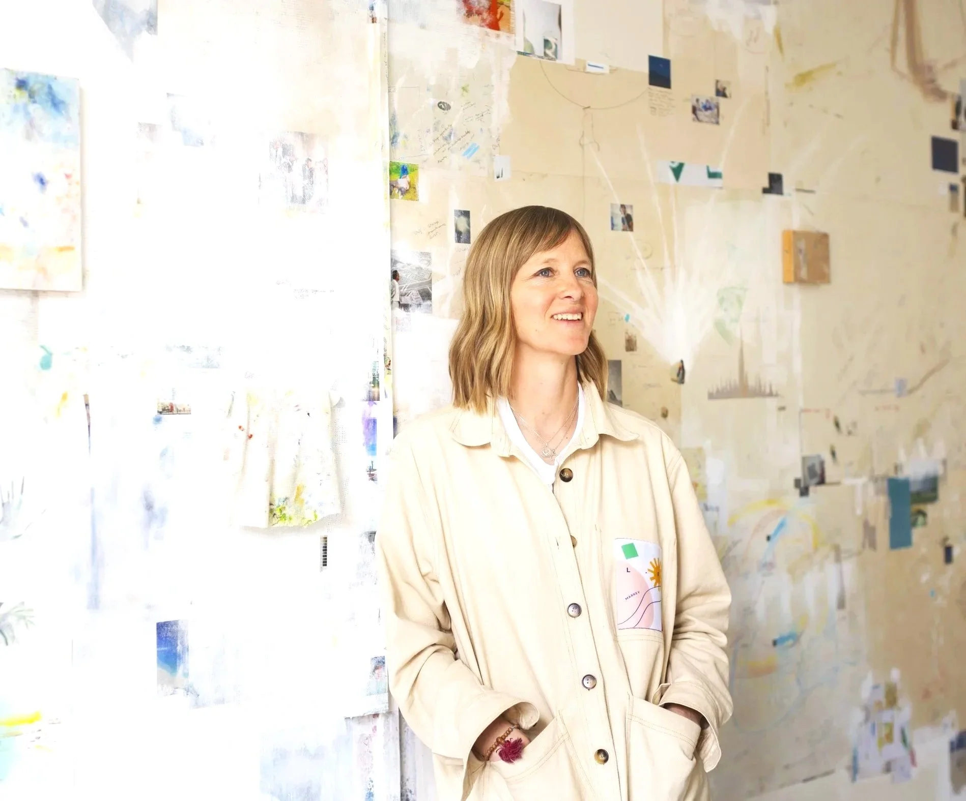 A woman with shoulder-length blonde hair smiling and looking to her right, standing in front of a wall decorated with various colorful papers, photos, and drawings.