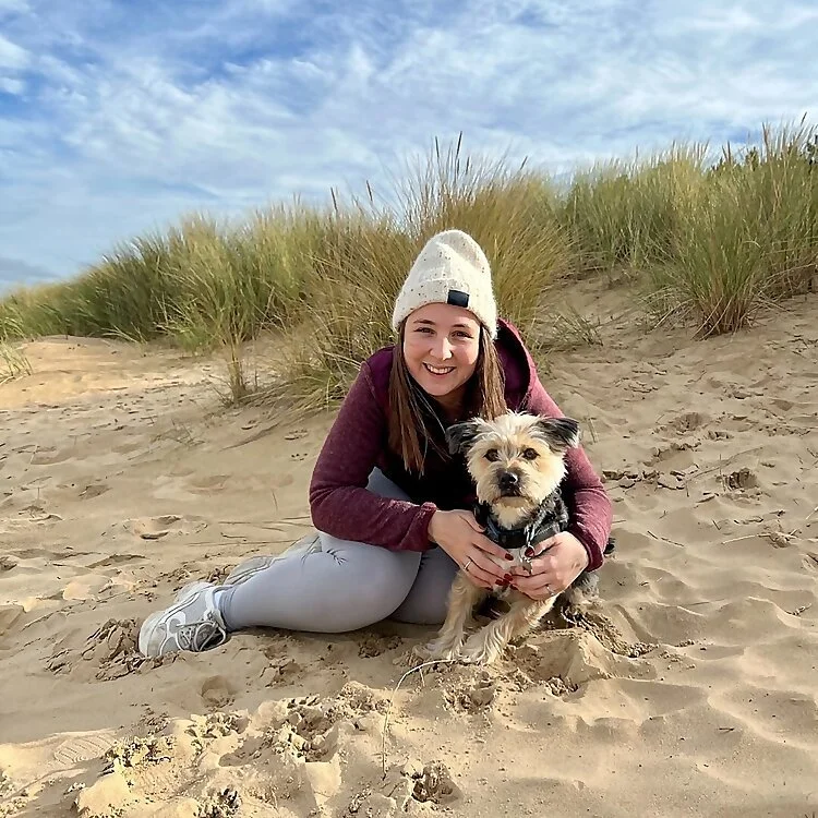 A young woman in a white beanie and purple jacket sitting on sandy beach with a small dog in her lap, with tall grass and a blue sky in the background.