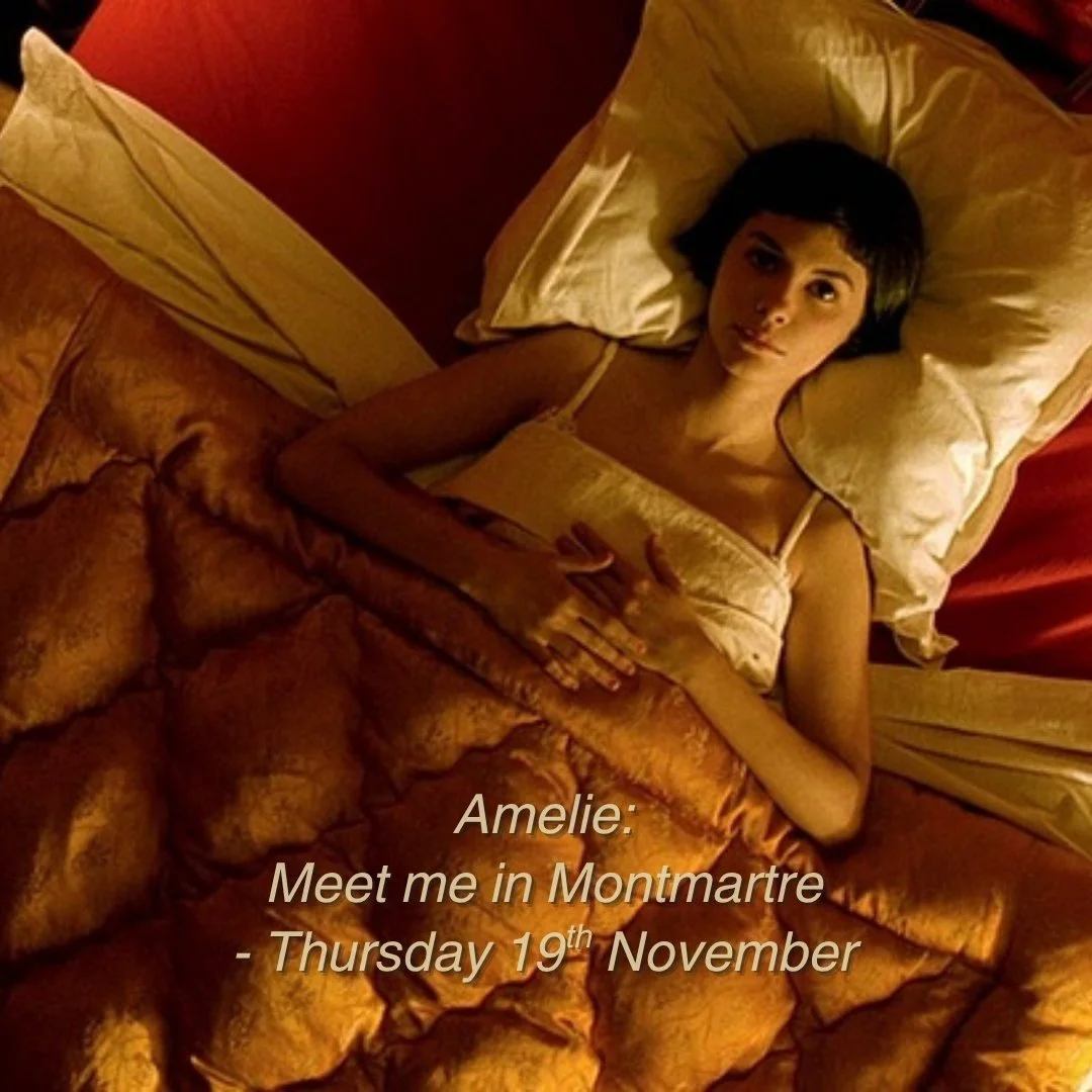 A woman with short black hair lying in bed with beige sheets and pillows, looking at the camera. There is a text overlay that reads: "Amelie: Meet me in Montmartre - Thursday 19th November."