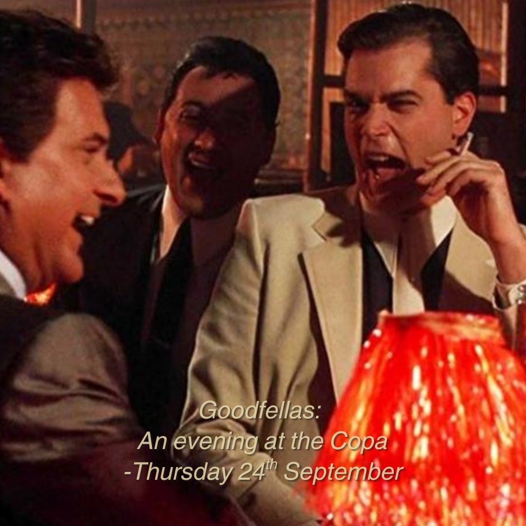 Three men in suits laughing and talking at a restaurant or bar, with warm lighting and a decorative lamp in the foreground.