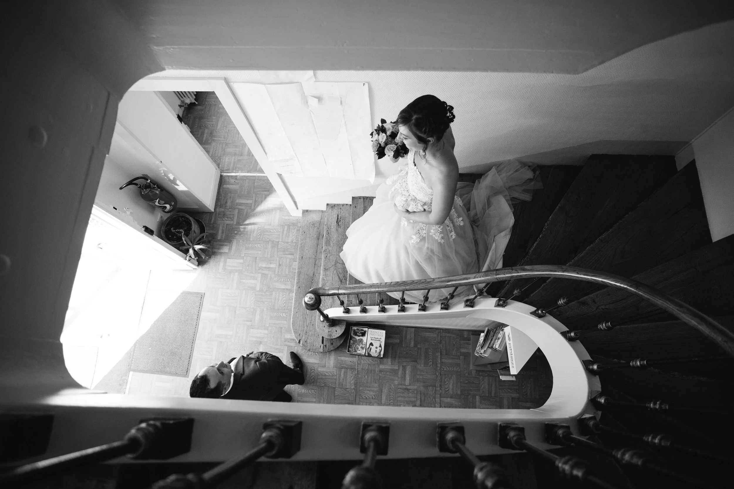 Une femme en robe de mariée tenant un bouquet de fleurs, vue du haut dans une maison classique avec escalier en bois.