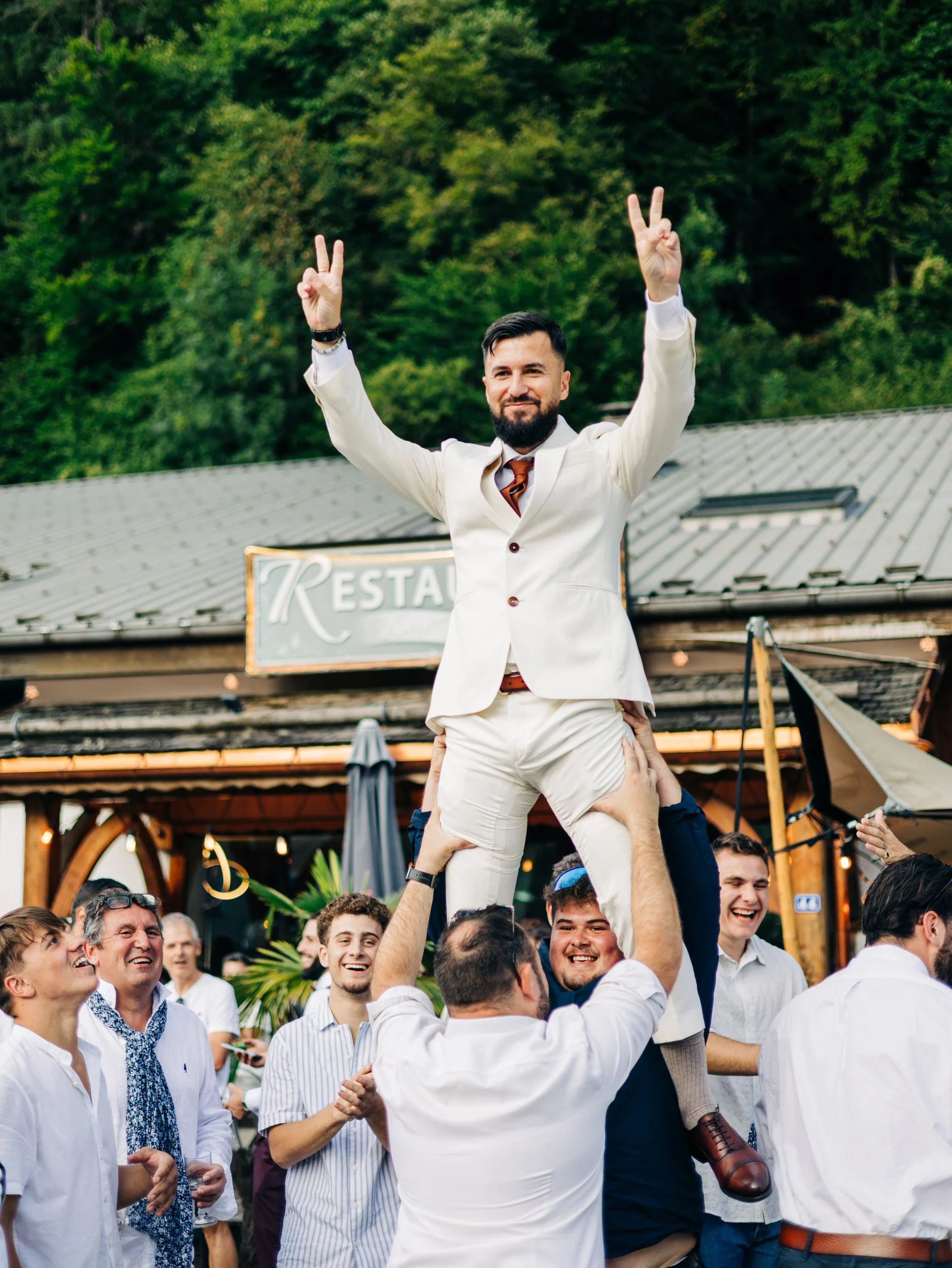 Un groupe de personnes souriantes lors d'une célébration en extérieur. Un homme en costume blanc, avec une barbe, est assis sur les épaules de plusieurs autres personnes, levant les deux mains en signe de paix. La scène montre une atmosphère joyeuse 