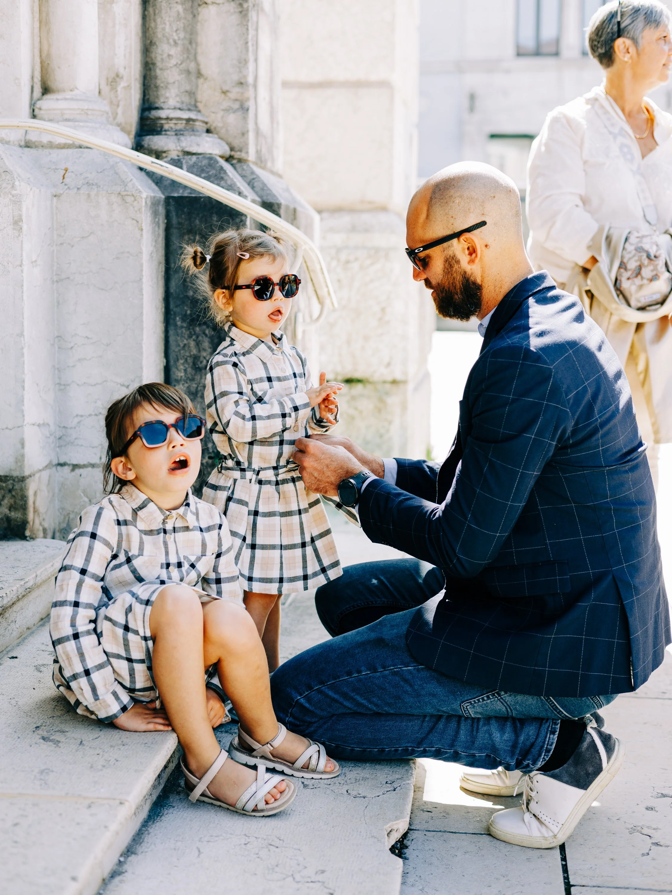 Un homme agenouillé donne une signature à une jeune fille qui porte des lunettes de soleil et une robe à carreaux, assise sur un escalier en pierre. Deux autres enfants, également portant des lunettes de soleil et des vêtements à carreaux, sont assis