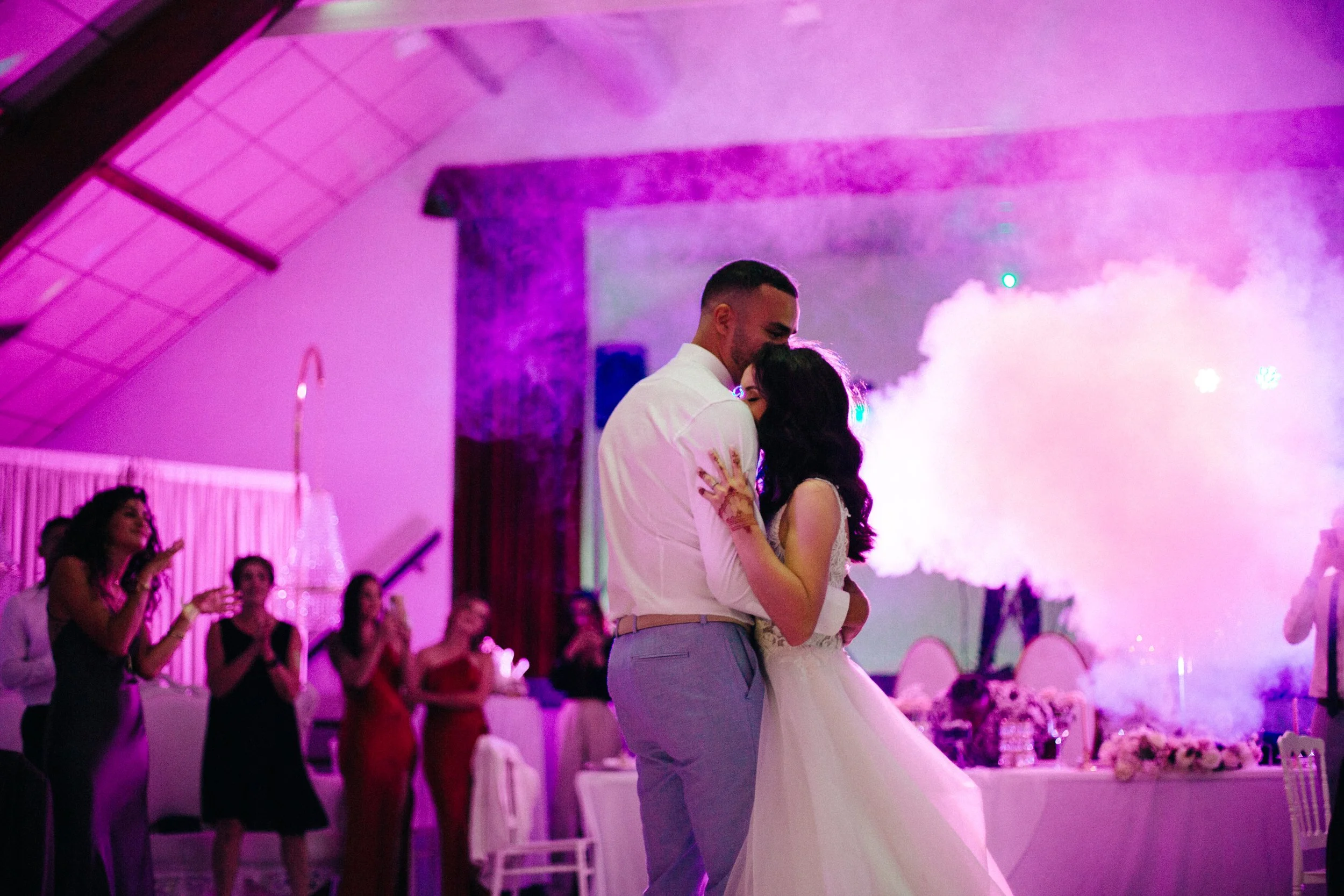Un couple en train de danser lors d'une cérémonie nuptiale dans une salle éclairée en violet. Il y a des invités qui regardent et prennent des photos, avec un décor floral en arrière-plan.