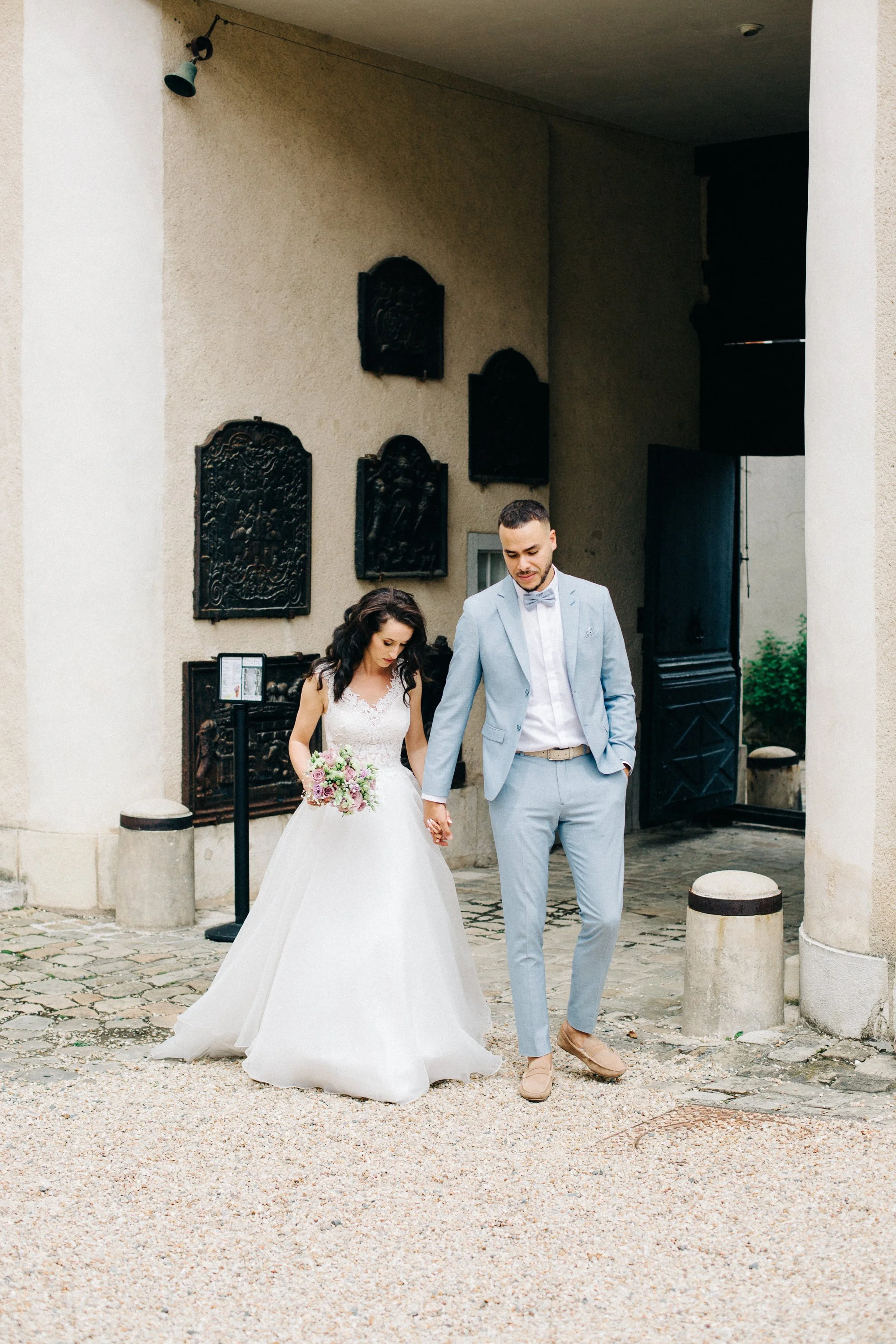 Un couple marié marche main dans la main devant un bâtiment en pierre, la femme porte une robe de mariage blanche et tient un bouquet de fleurs, l'homme porte un costume gris clair et une chemise blanche