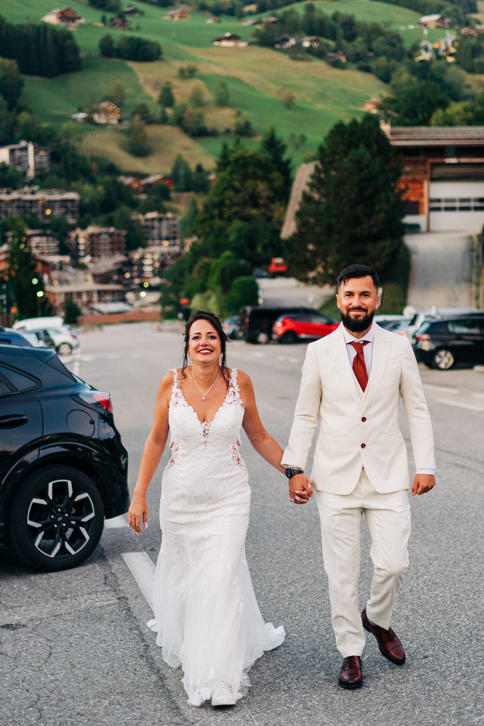 Un couple de mariés marche dans un parking, la femme porte une robe de mariée blanche et le homme un costume blanc avec une cravate rouge, ils se tiennent la main et sourient.