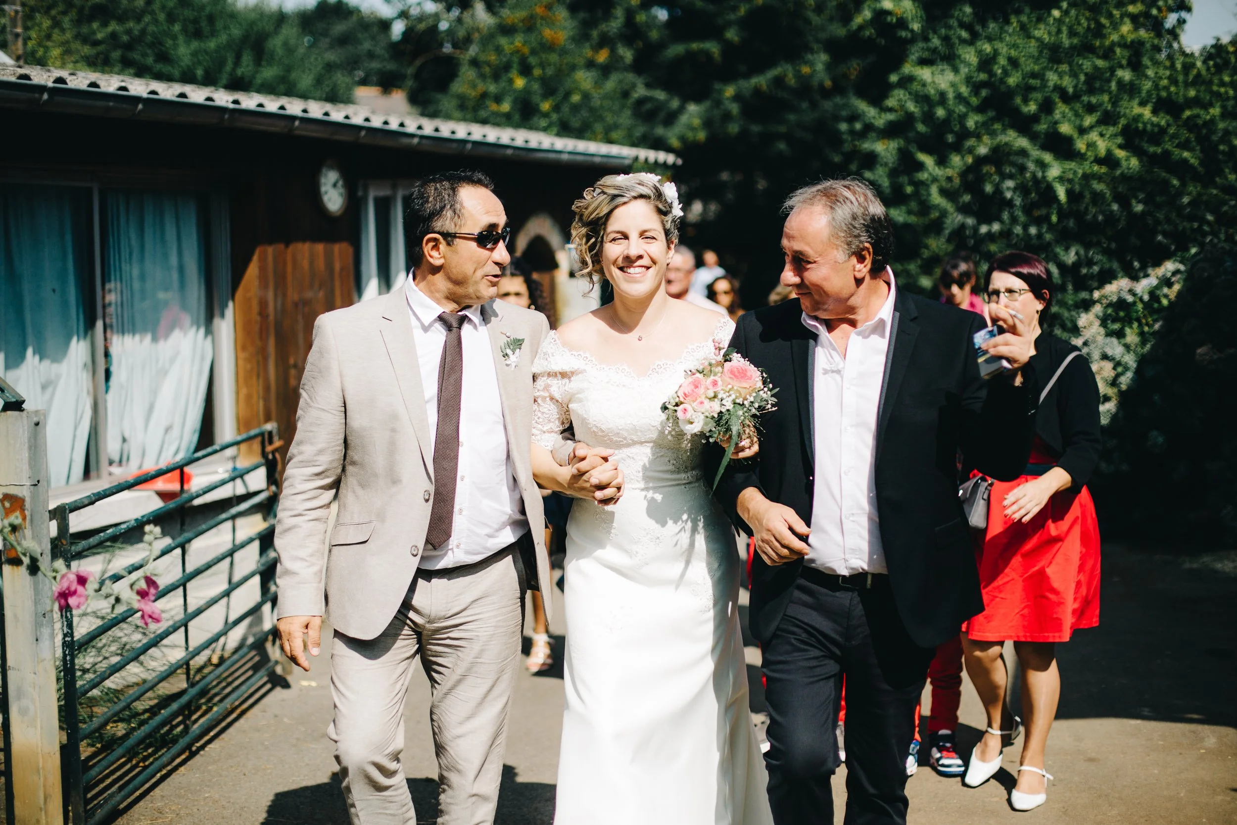 Un groupe de personnes lors d'un mariage en extérieur, dont une mariée souriante portant une robe blanche et tenant un bouquet de fleurs roses, entourée de deux hommes en costume. D'autres invités en arrière-plan.