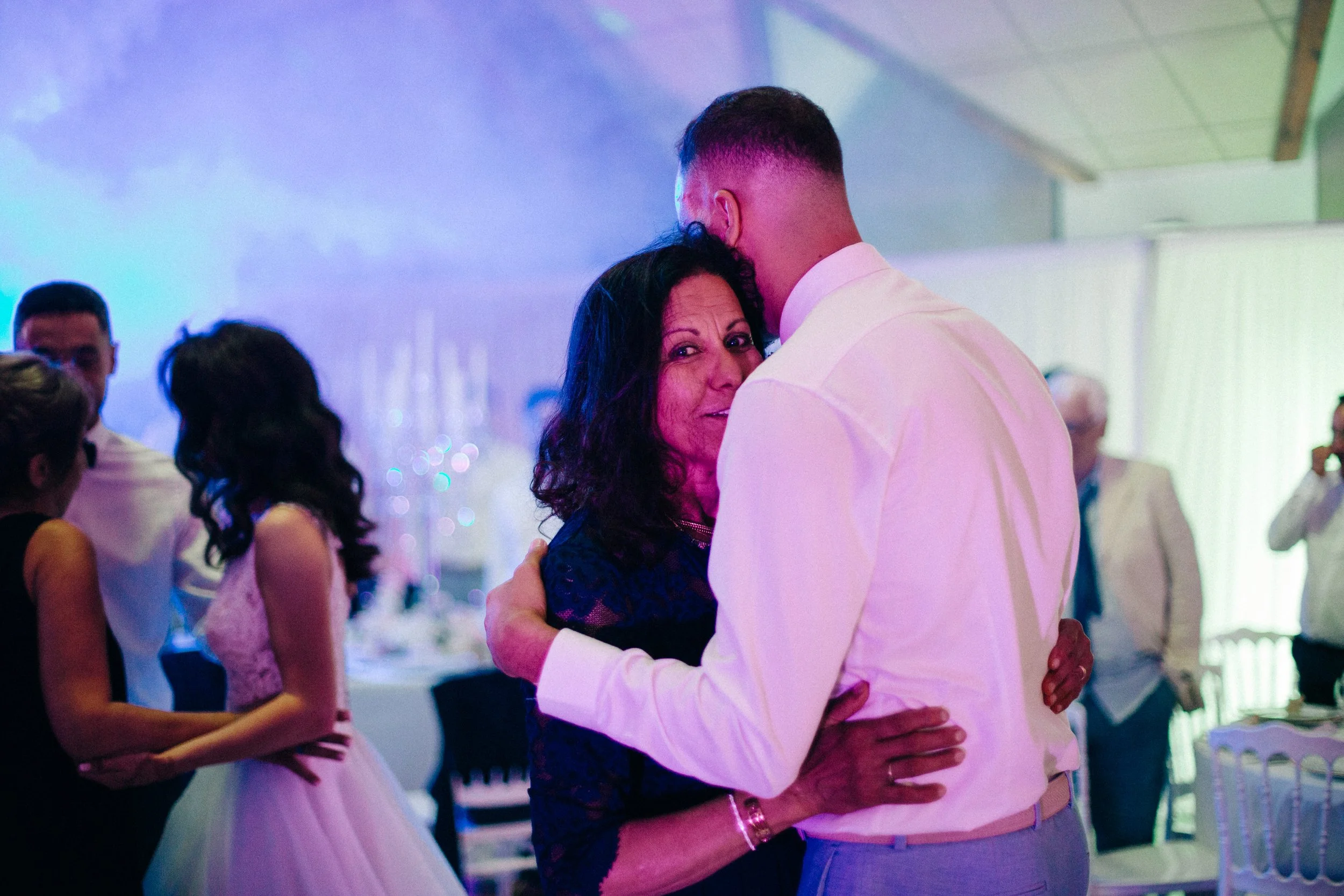 Une femme et un homme dansent étroitement lors d'une fête ou réception, la femme regarde vers la caméra avec une expression chaleureuse.